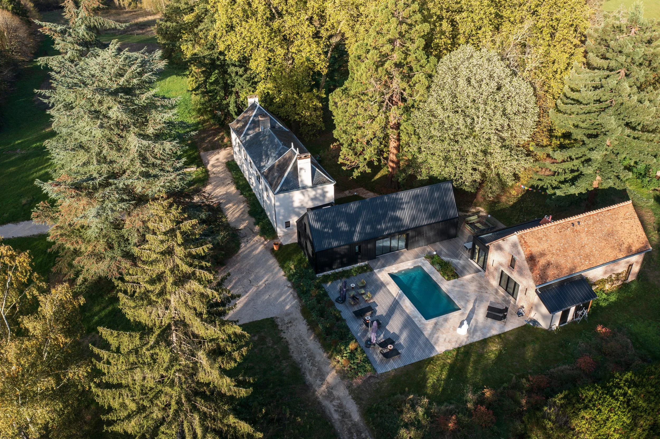 Aerial view of a backyard with a swimming pool, outdoor furniture, and a black modern building surrounded by large trees and a mix of houses and green landscape.