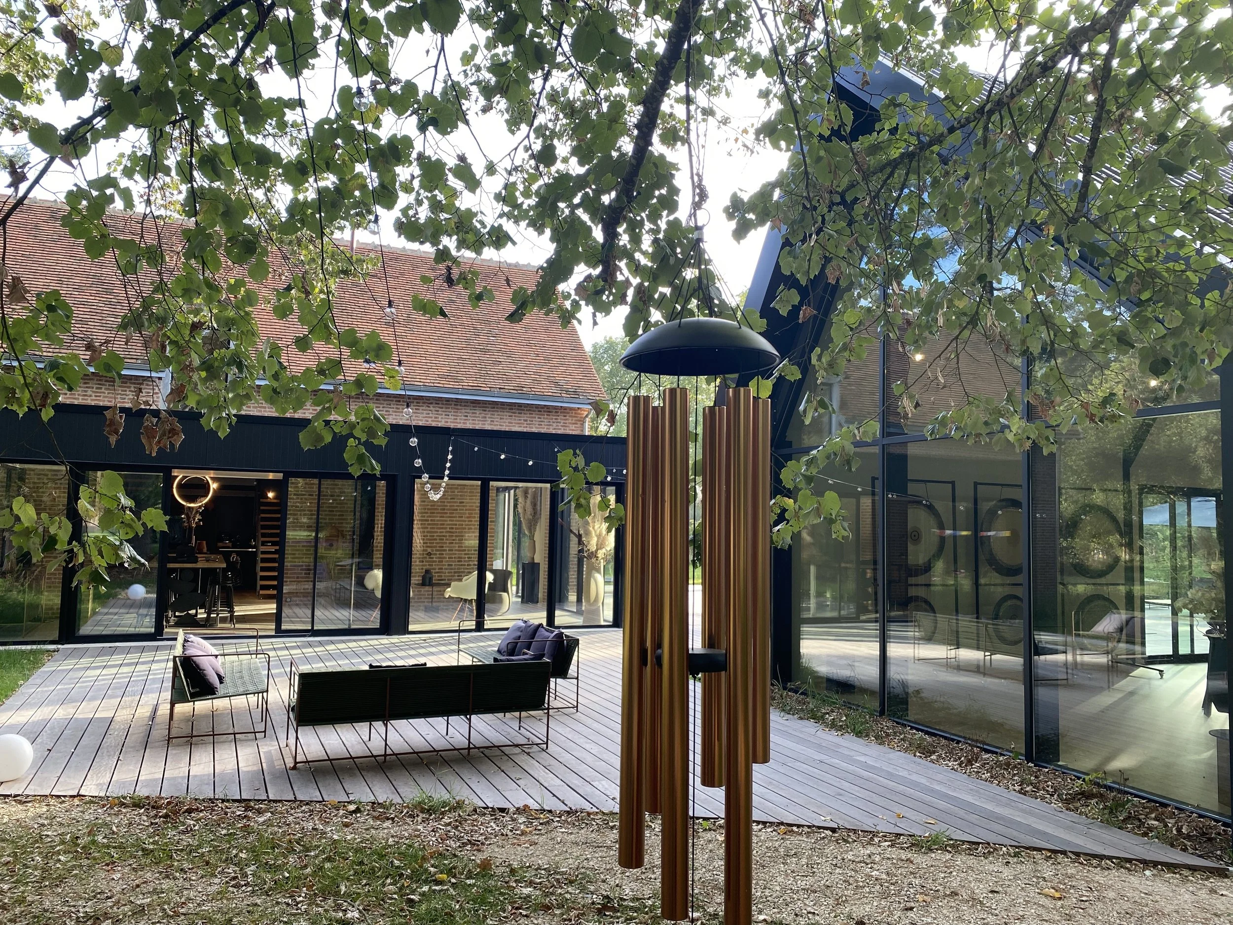Outdoor patio area with seating and a wind chime, surrounded by trees and a modern glass building.