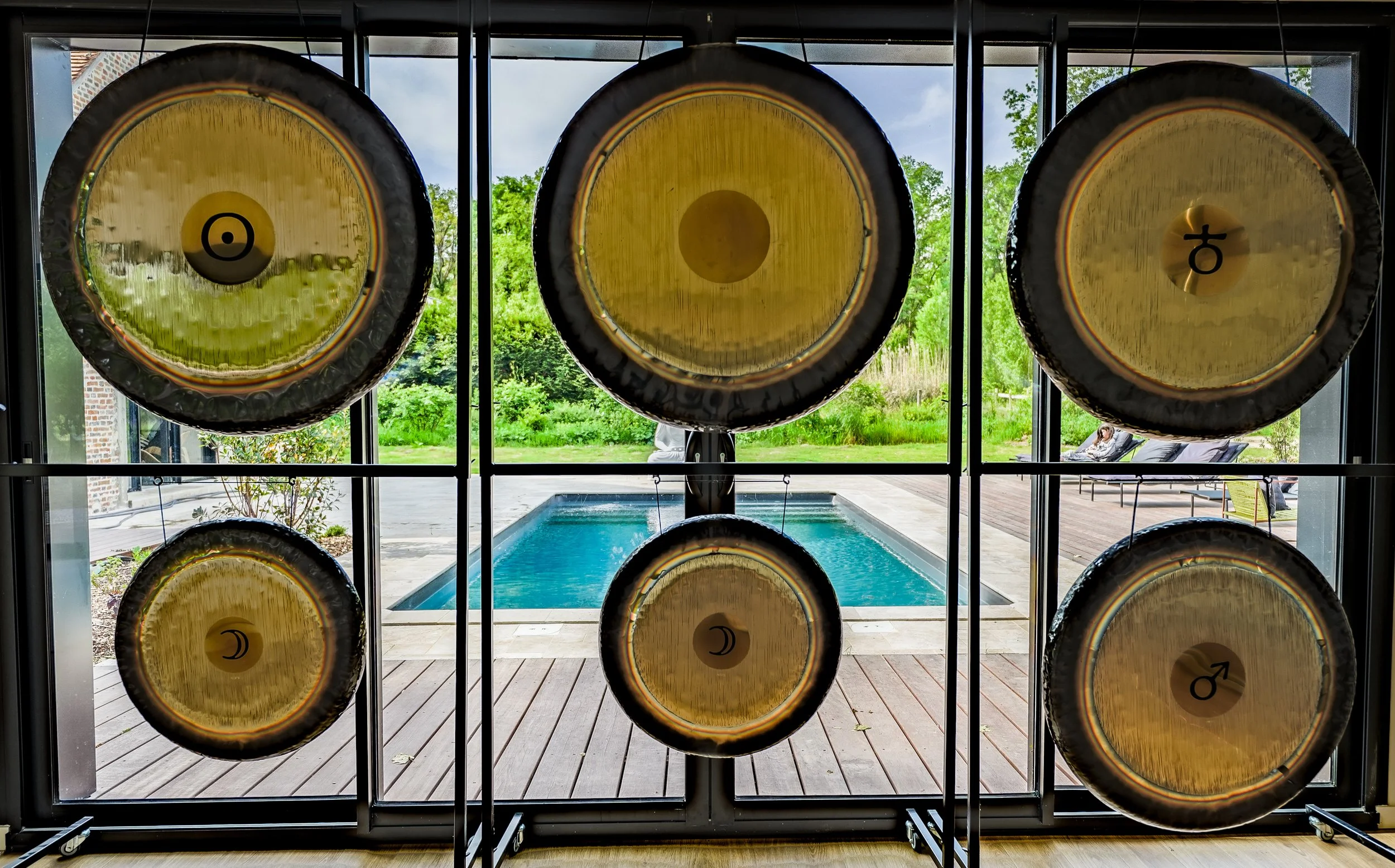 Six large gong drums mounted on a metal frame with a pool and greenery outside in the background.