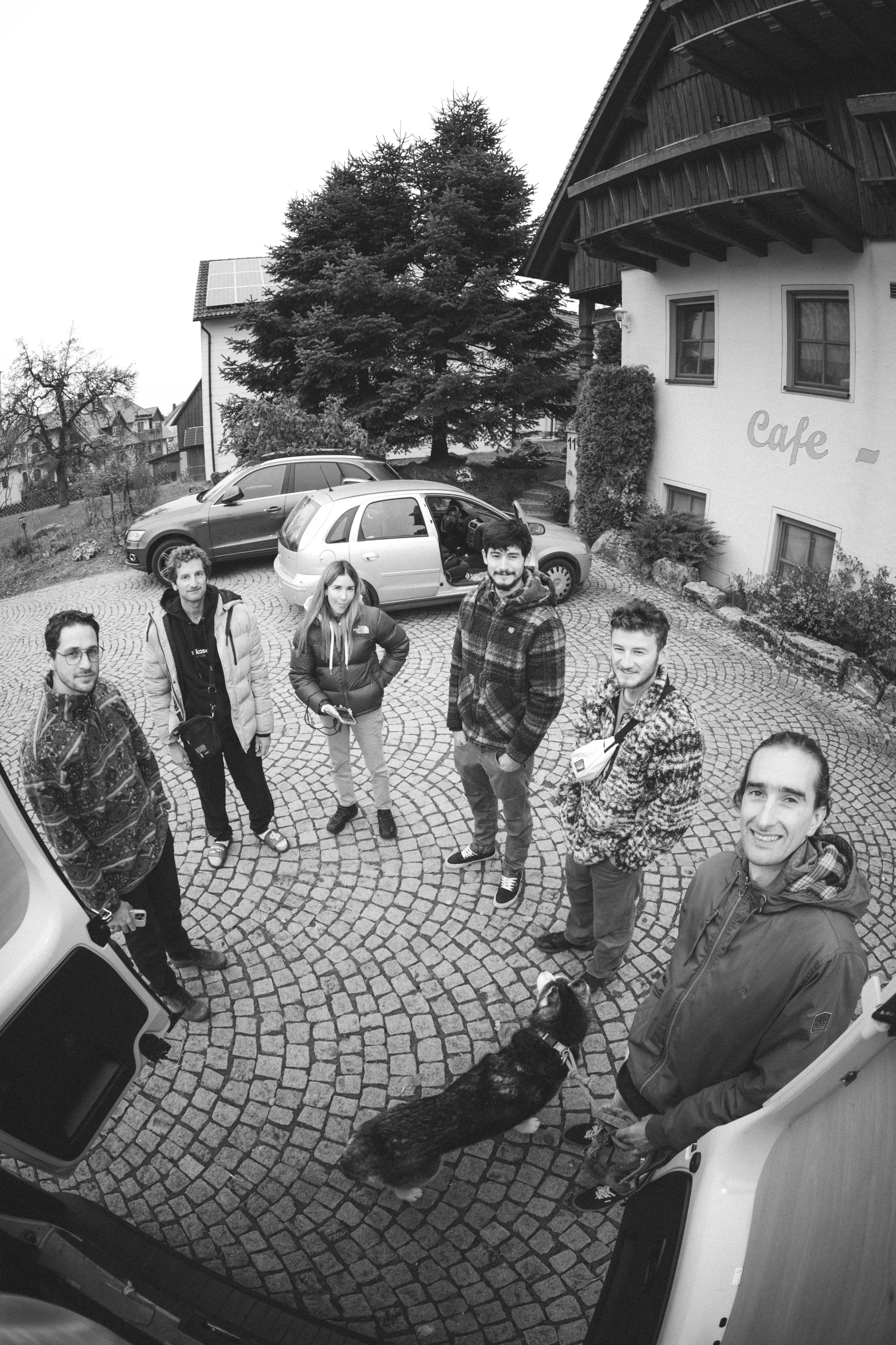 Groupe de six personnes devant un bâtiment avec un chien, en extérieur sur une place pavée.