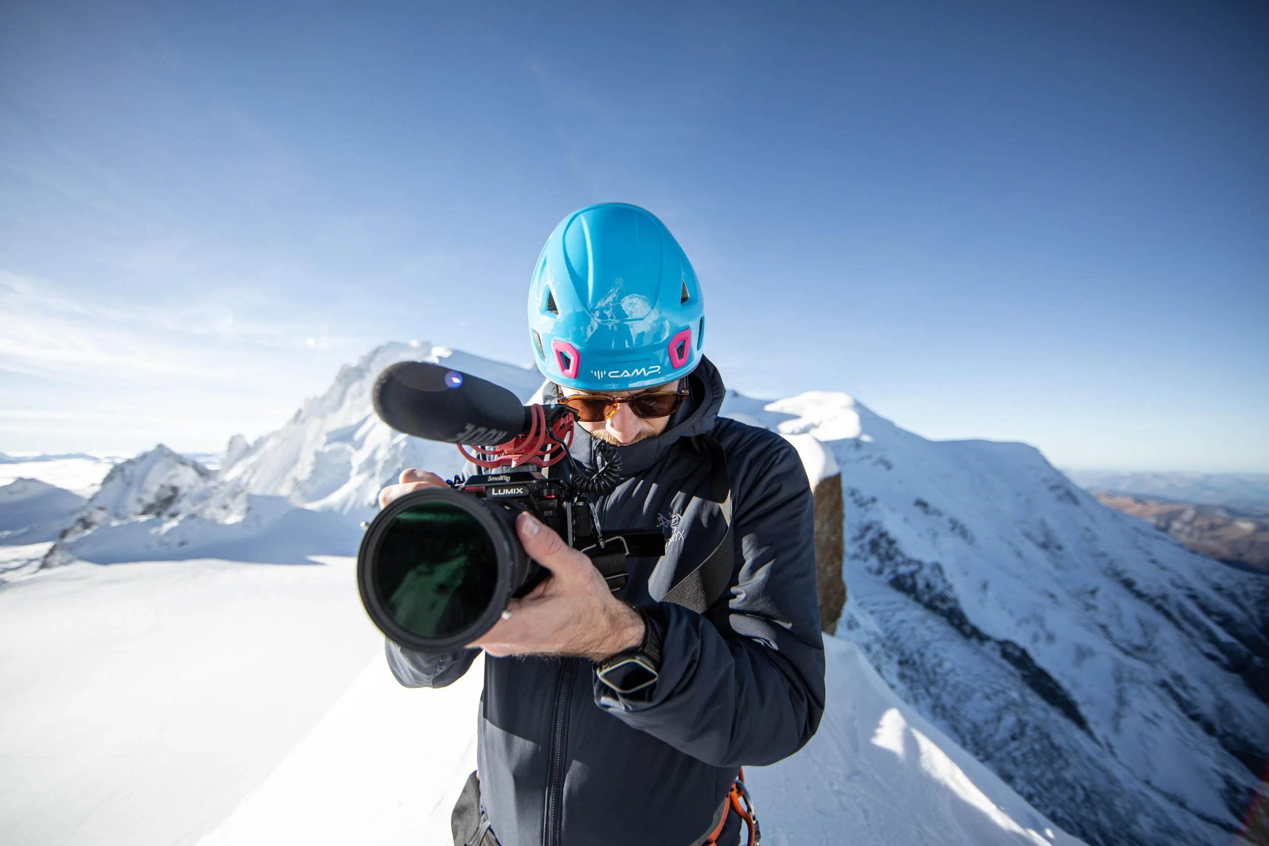 Un homme avec un casque bleu et des lunettes de soleil tient une caméra en pleine montagne enneigée.