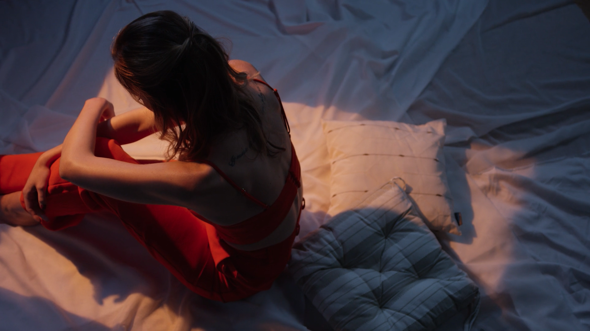 Une femme assise sur un lit avec des coussins, vue de dessus, portant une robe rouge/orange, dans une ambiance tamisée.