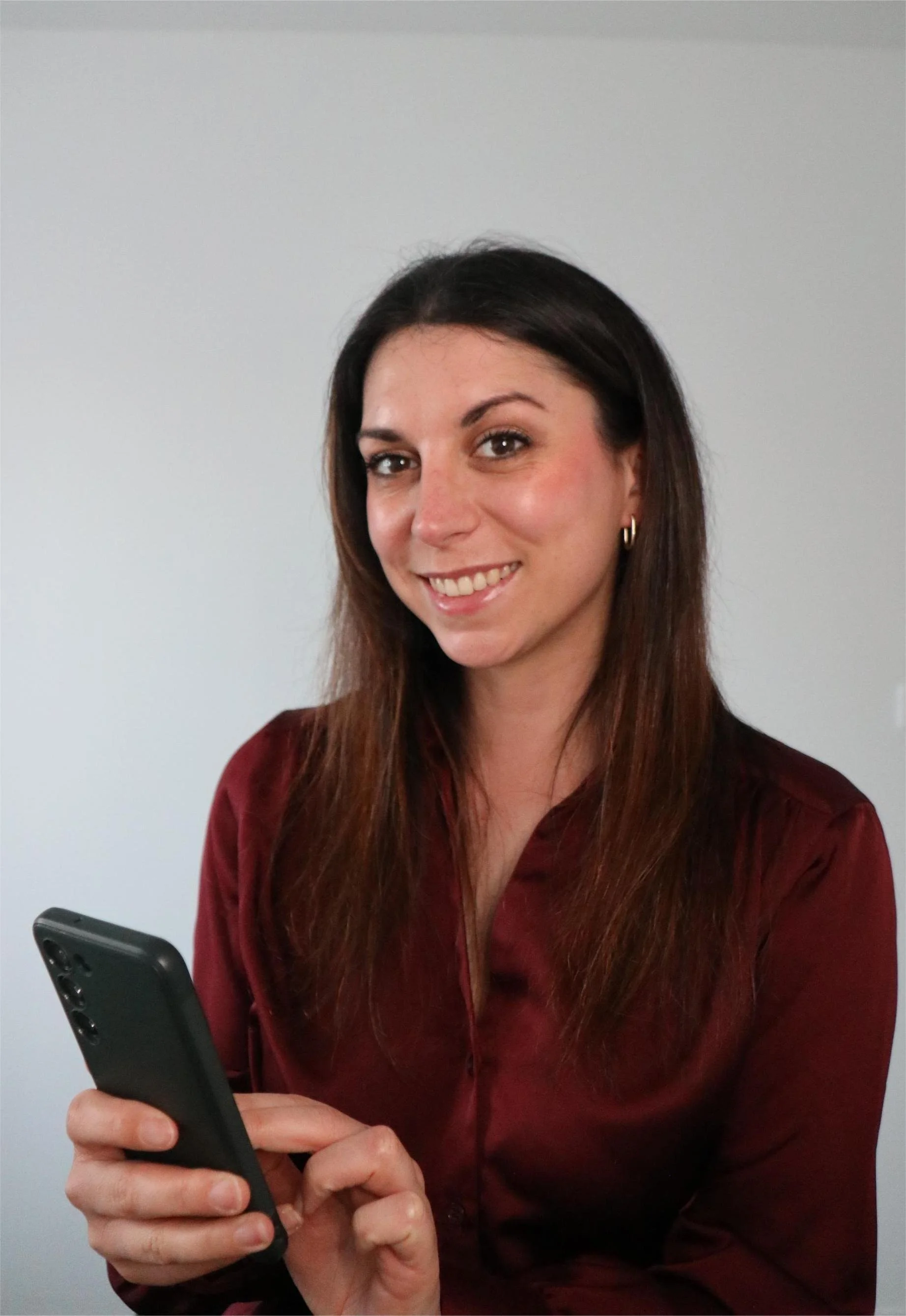 Jeune femme souriante avec boucle d'oreille en or, tenant un smartphone noir, portant une chemise rouge en satin, fond blanc.