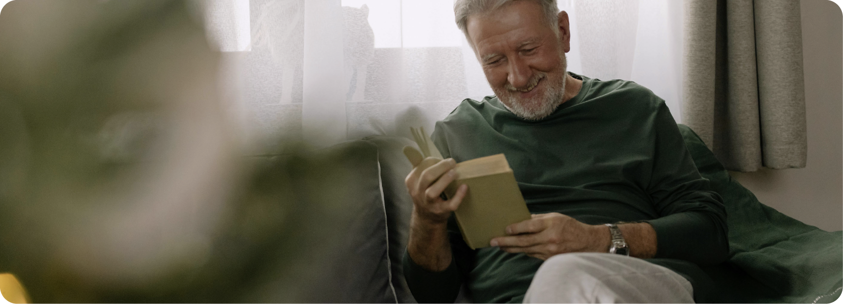 Un homme âgé souriant assis sur un canapé, lisant un livre dans une pièce lumineuse avec des rideaux blancs.