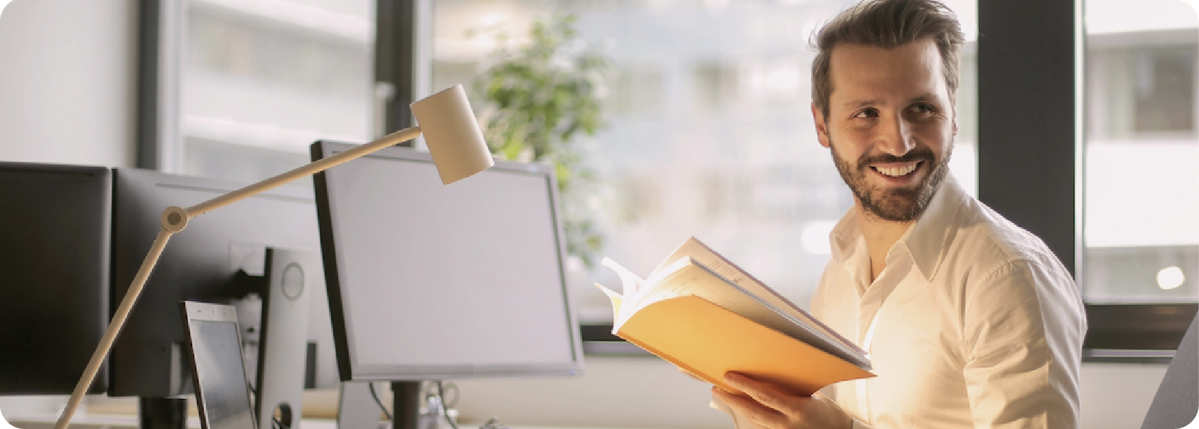 Un homme souriant tenant un livre ouvert dans un bureau lumineux avec plusieurs écrans d'ordinateur et une lampe de bureau.