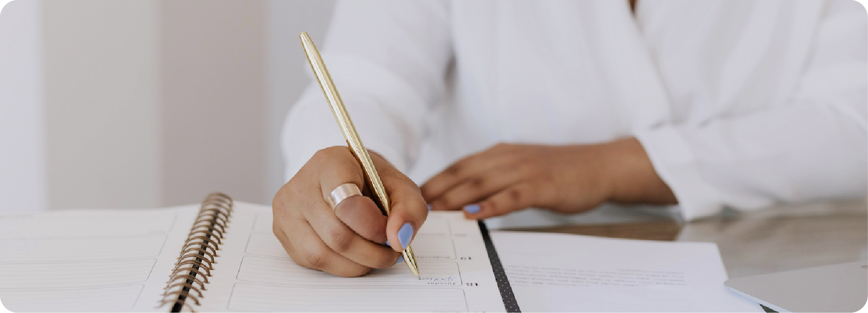 Une personne écrit dans un carnet avec un stylo doré, avec un autre document à côté.