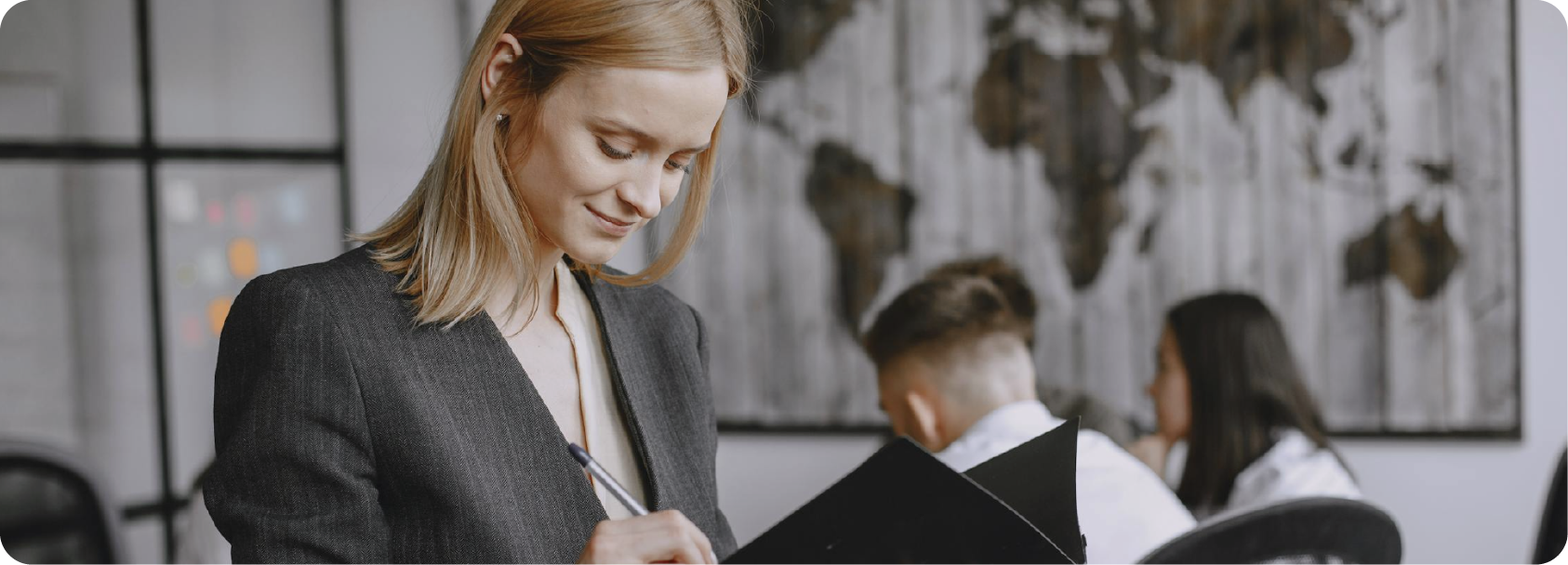 Une femme souriante écrit dans un carnet dans un espace de travail collaboratif avec d'autres personnes à l'arrière-plan.