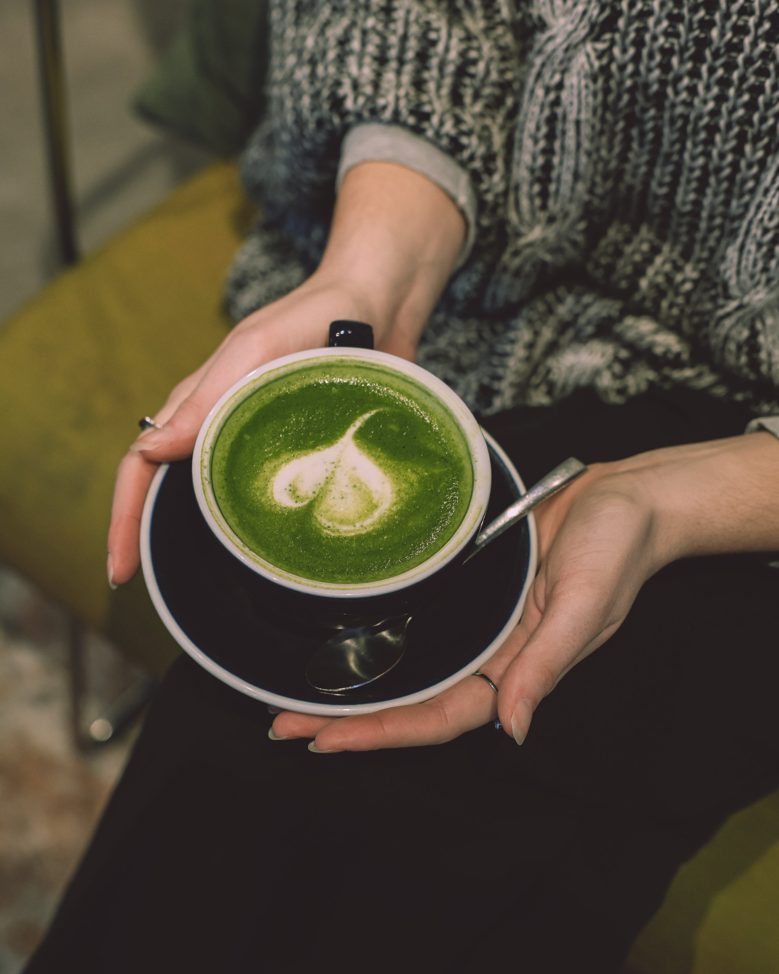 Person holding a black cup of matcha latte with latte art, sitting on a yellow cushion.