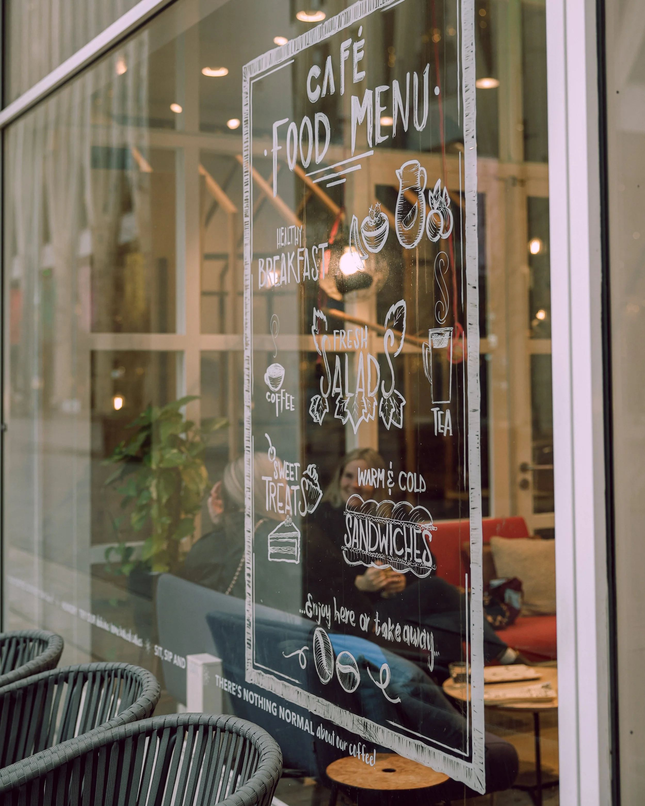 Glass window with a hand-painted menu for a cafe, listing food and beverage options including breakfast, salads, sandwiches, coffee, tea, and desserts, with some illustrations of food and drinks.