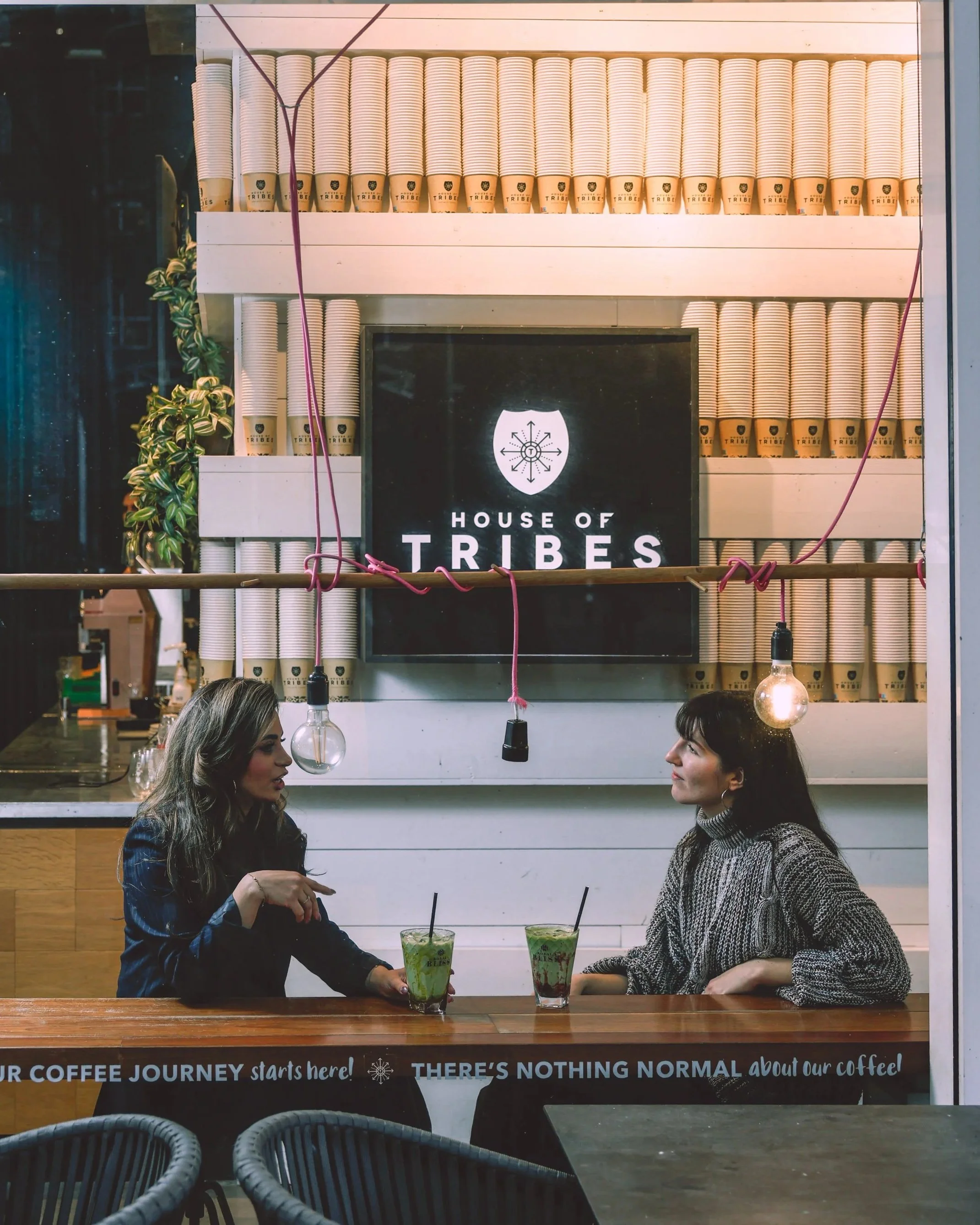 Two women sitting at a wooden counter in a coffee shop, engaged in conversation with drinks in front of them, with a sign saying 'House of Tribes' on the wall behind them, surrounded by stacked disposable coffee cups and minimalistic lighting.