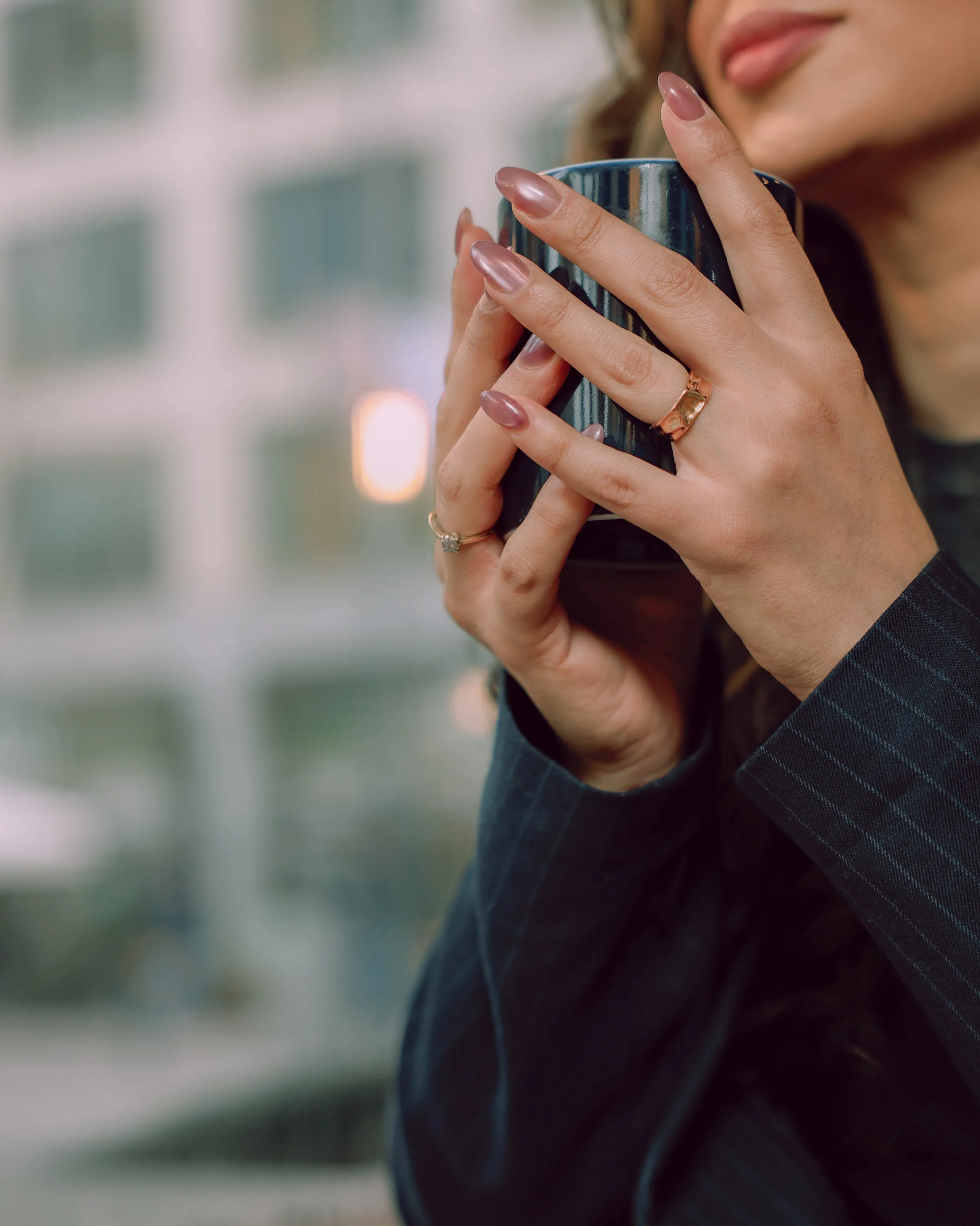 Close-up of a woman holding a dark mug, with her fingers adorned with rings, while wearing a pinstripe blazer.