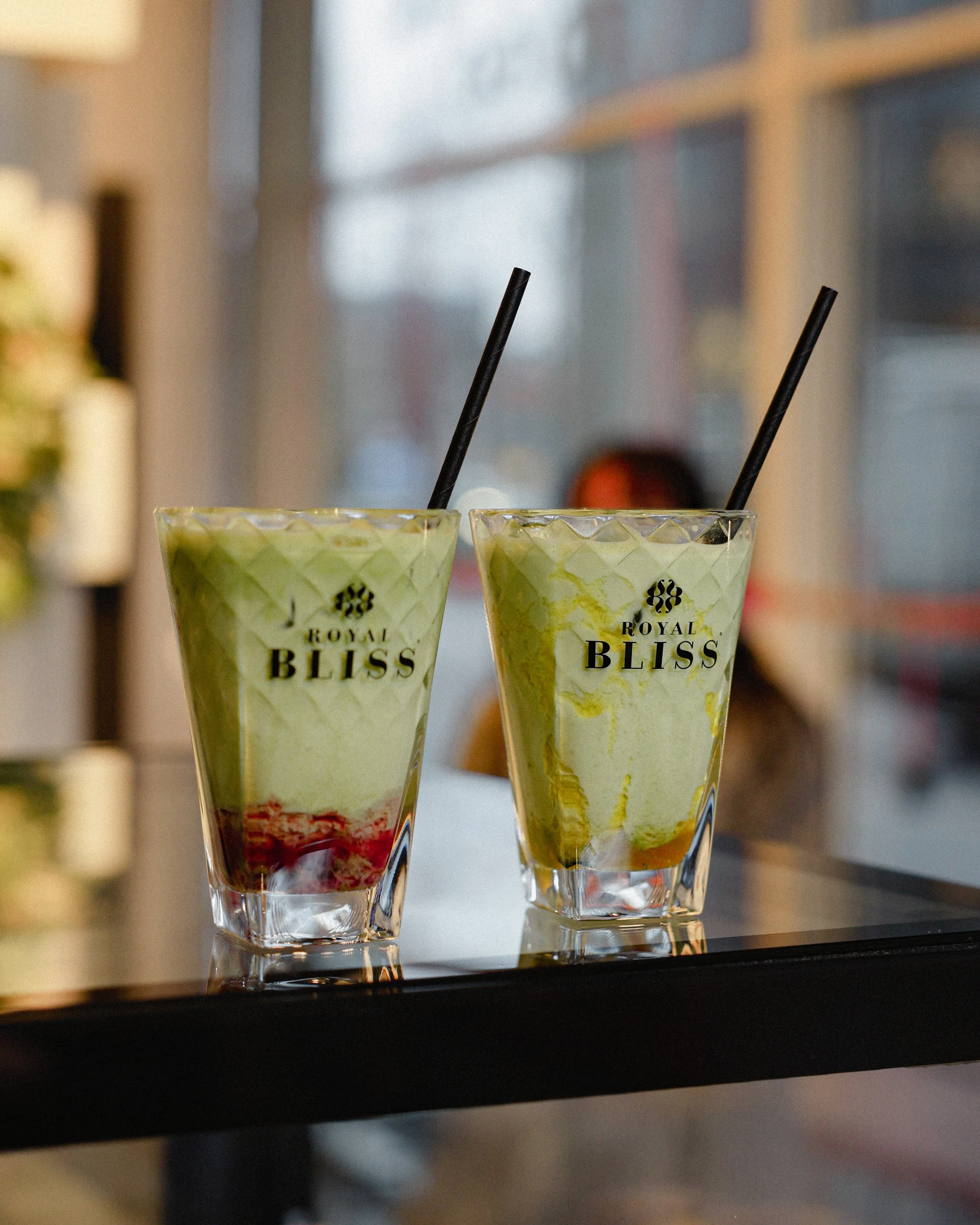 Two glasses of green smoothie with black straws on a black counter, with a blurred background and a 'Royal Bliss' logo on the glasses.