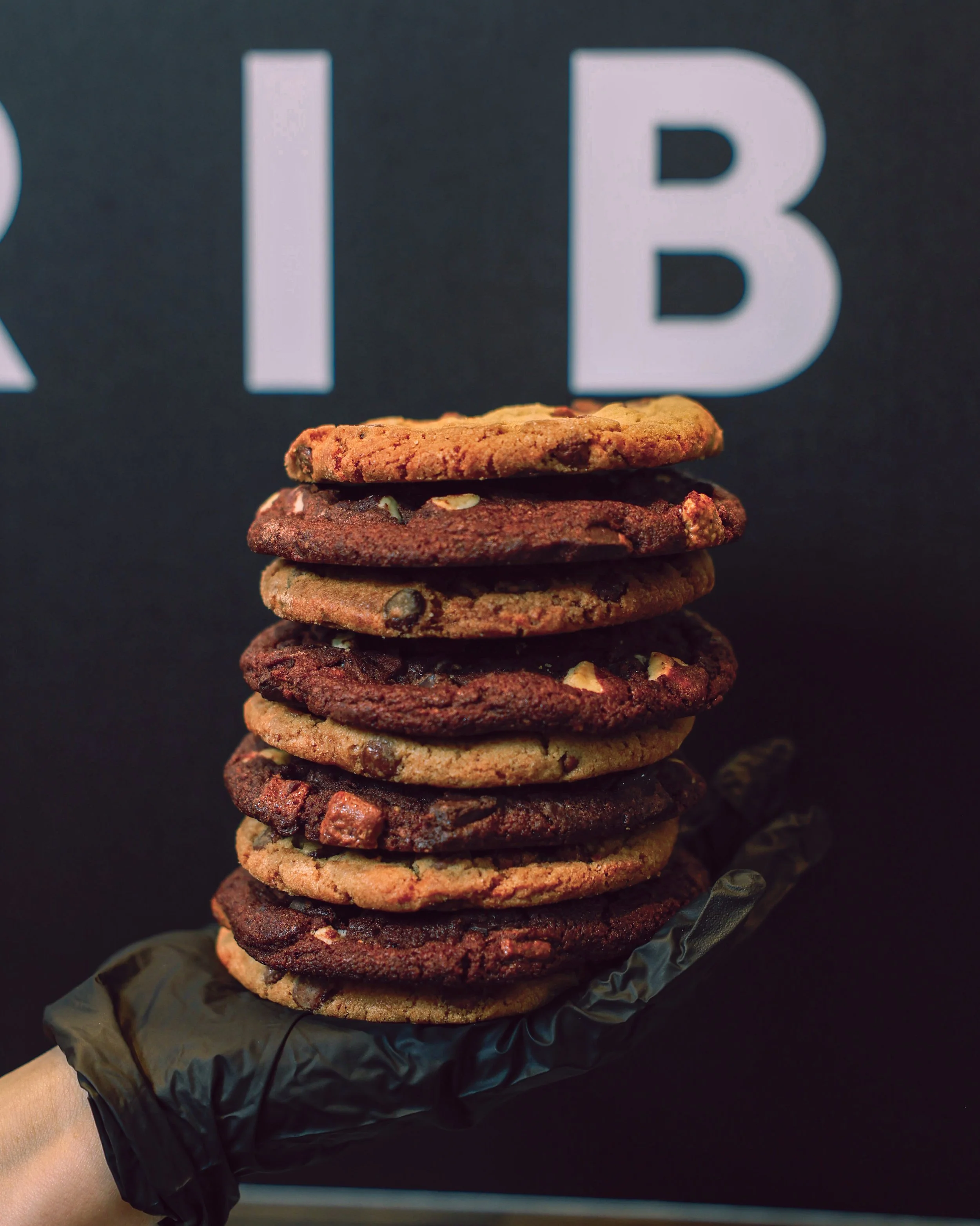 A hand wearing a black glove holds a stack of several cookies in front of a black background with large white letters.