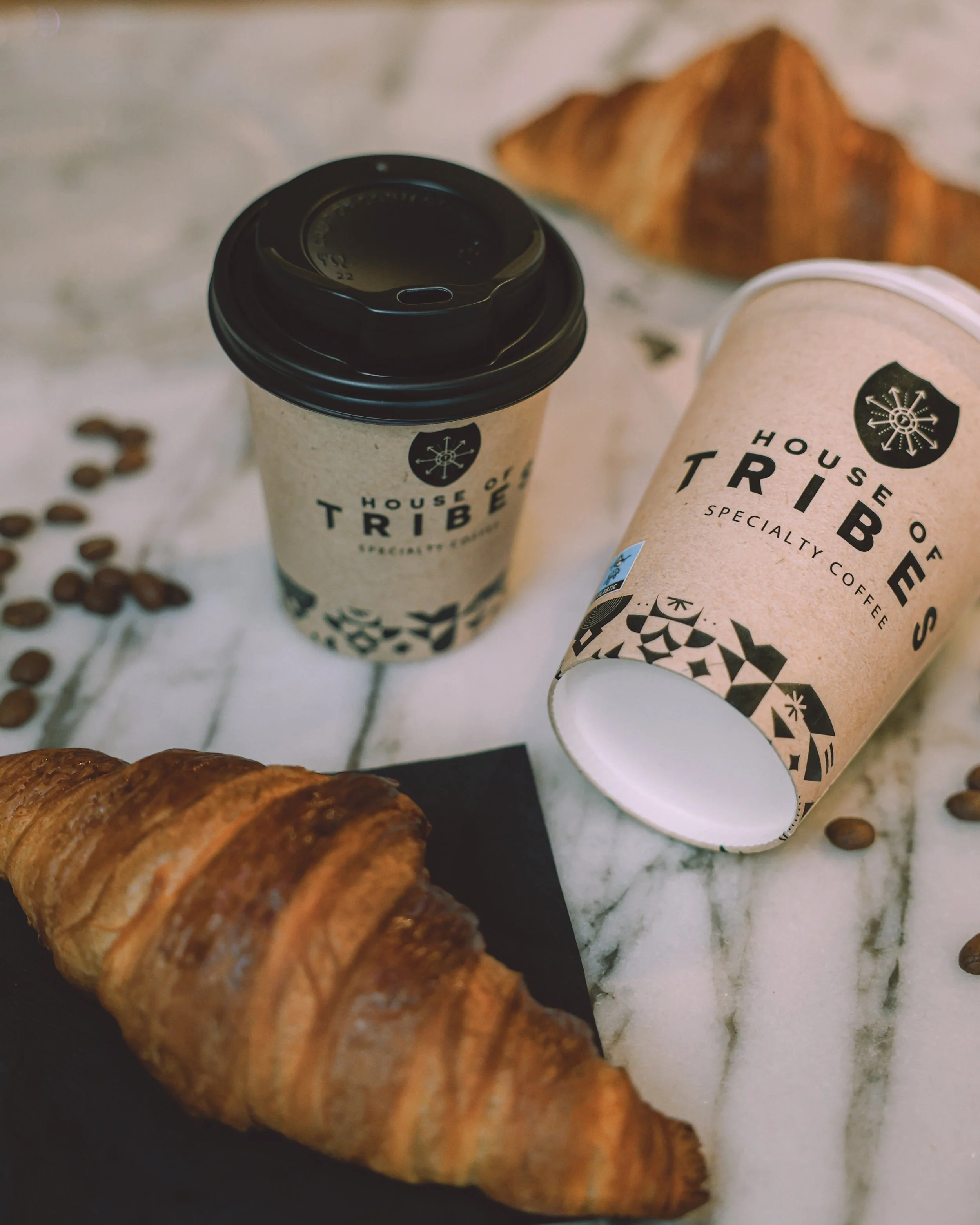 Two disposable coffee cups with the label 'House of Tribes Specialty Coffee,' surrounded by croissants, scattered coffee beans, on a white marble surface.