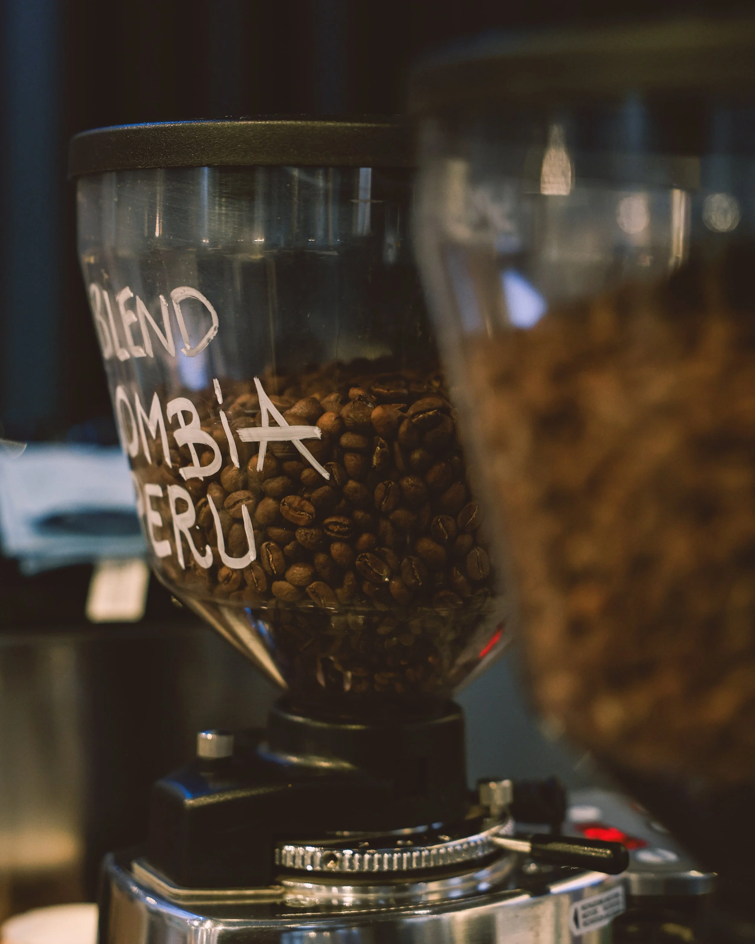 Close-up of a coffee grinder filled with coffee beans, with handwritten labels on the glass container that read "BLEND" and "ARABICA".