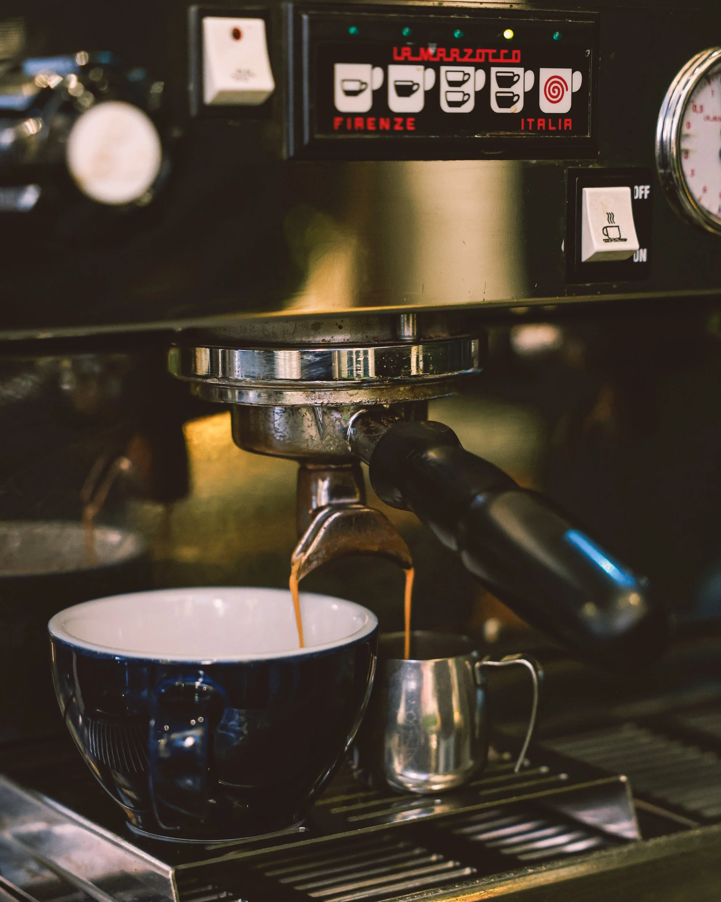 Close-up of an espresso machine dispensing coffee into a black and white mug, with a small metallic pitcher nearby.