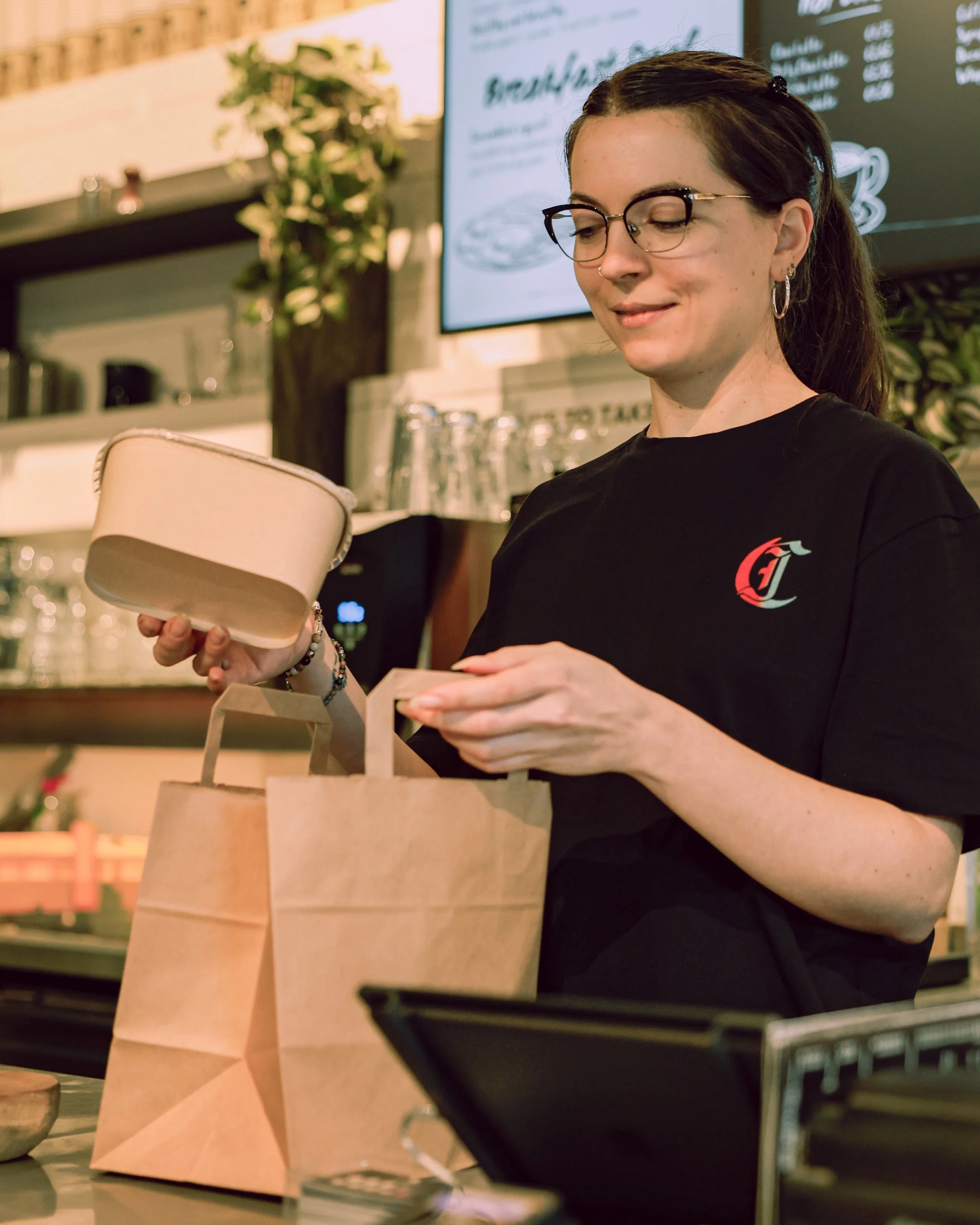 A woman with glasses and dark hair in a ponytail is opening a paper bag at a cafe counter. She is wearing a black T-shirt with a logo on it, holding a small beige container in her right hand, with a brown paper bag in front of her.