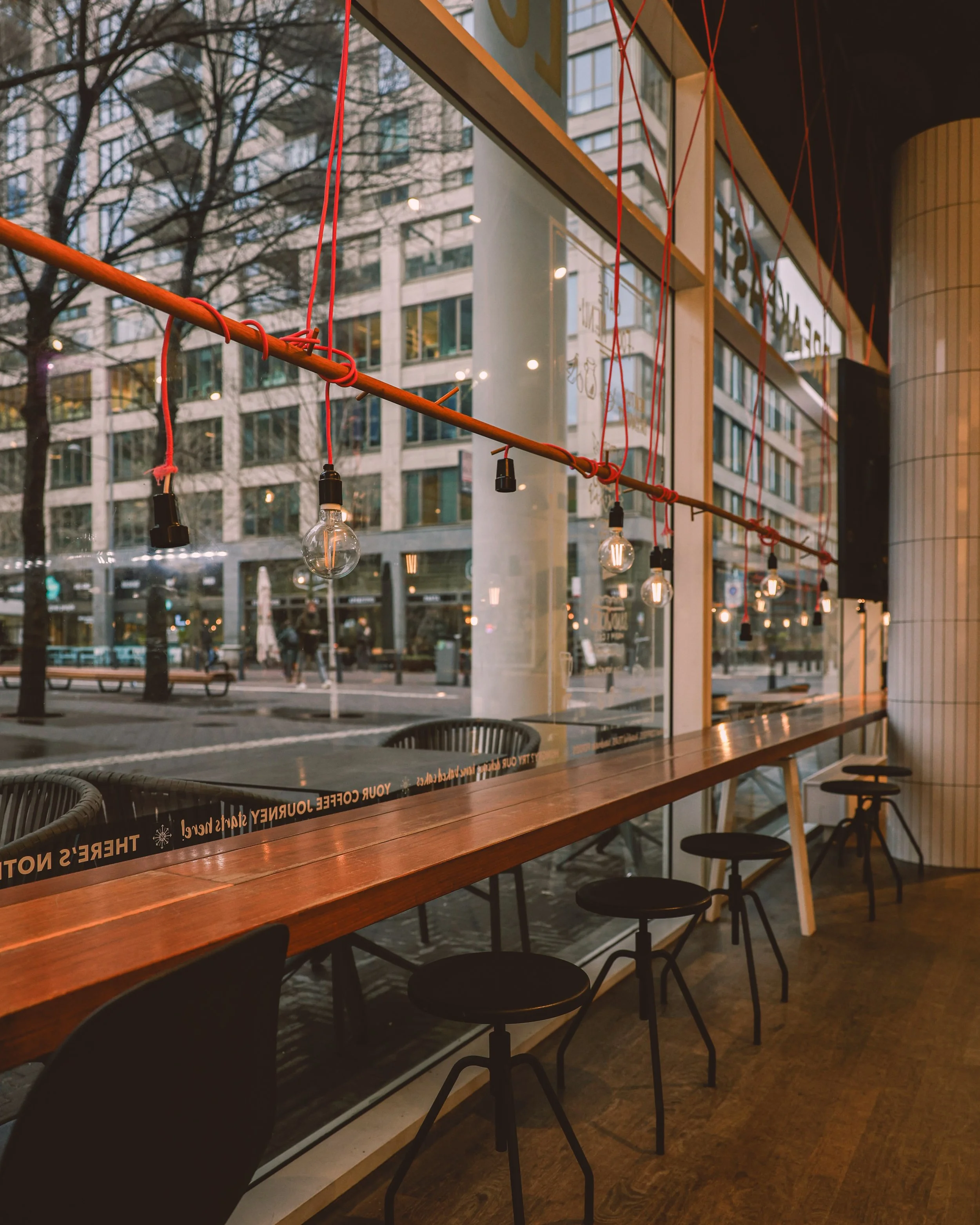 Interior of a modern cafe with a wooden counter and black bar stools near large windows. Red string lights with exposed bulbs hanging across the window. Cityscape with buildings and trees outside.