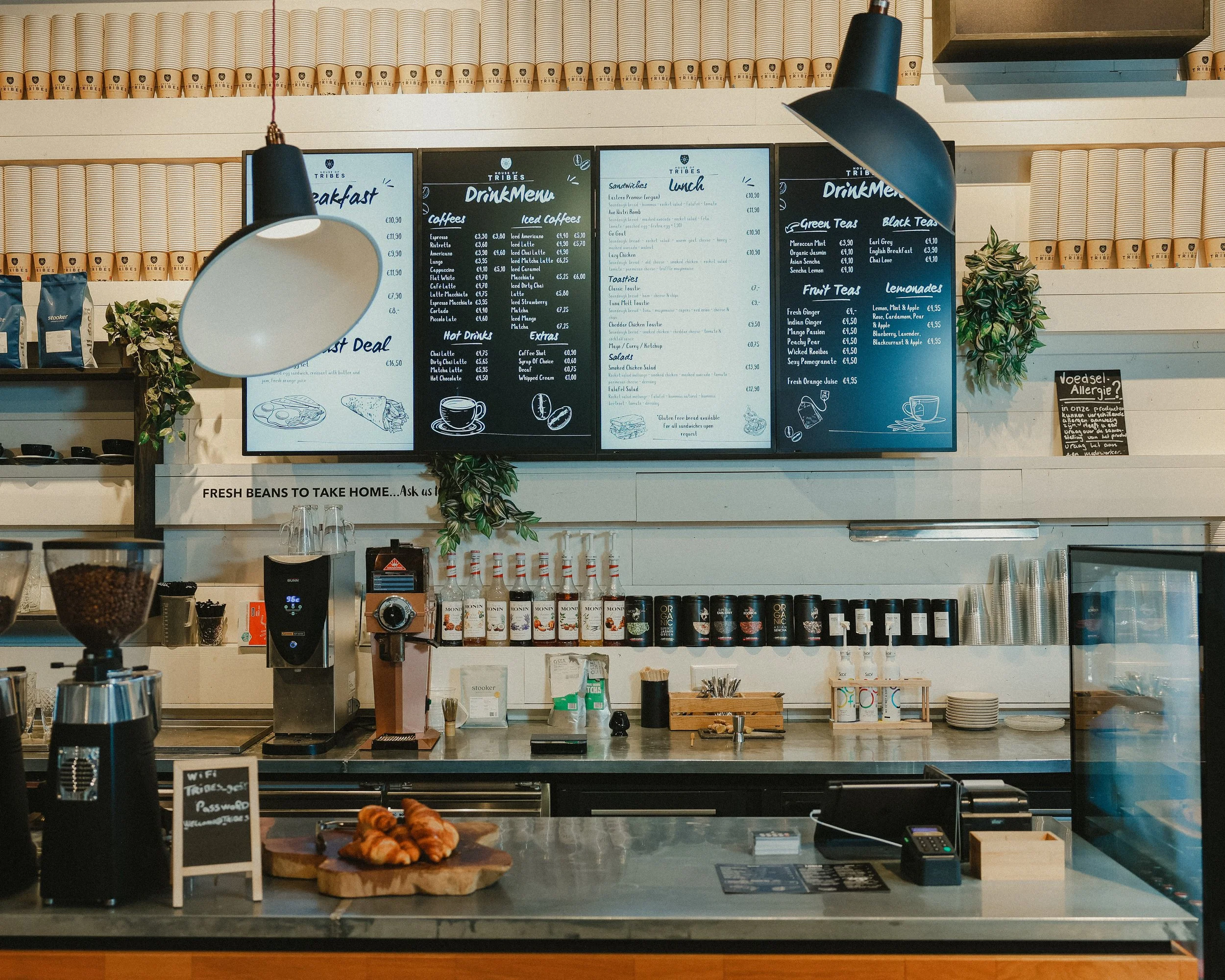 Interior of a coffee shop with black menu boards, coffee equipment, and pastries on the counter, decorated with plants and hanging lights.
