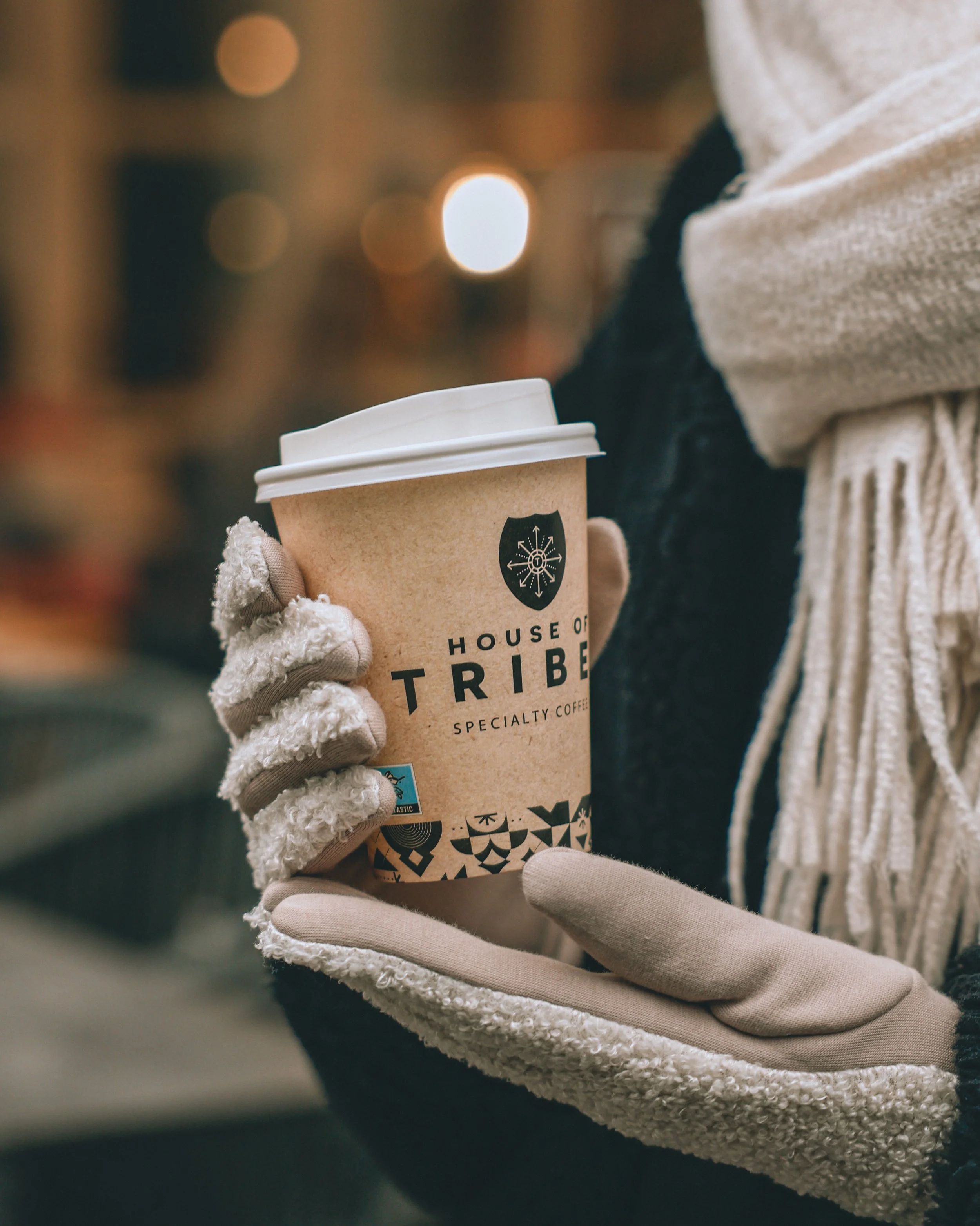 Person wearing gloves holding a paper cup of coffee with a lid at a coffee shop.