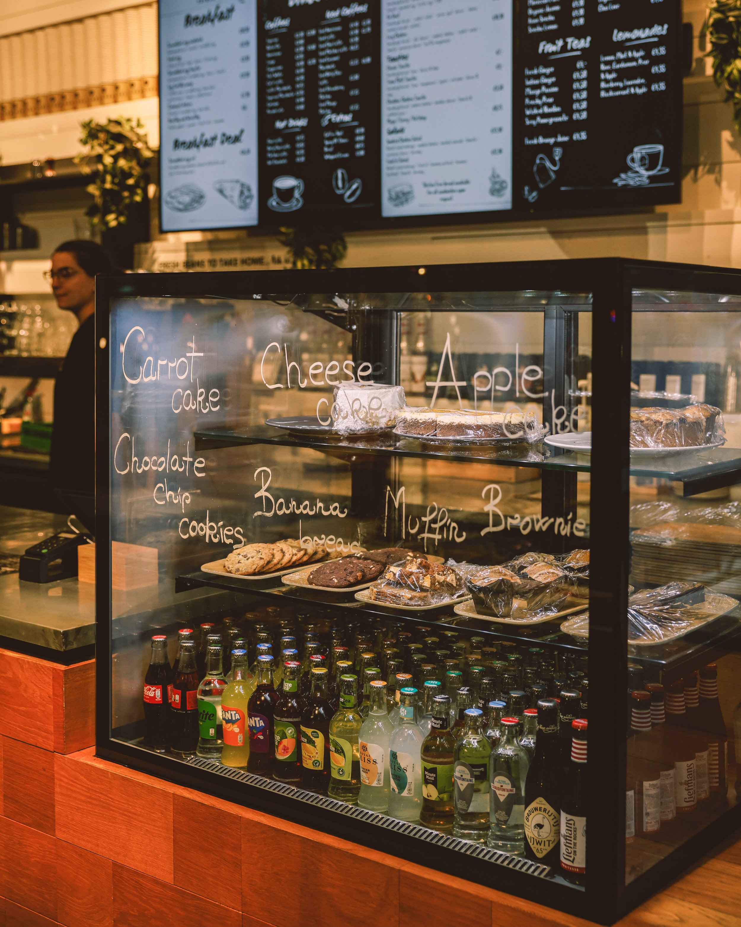 Glass display case with baked goods labeled as carrot cake, cheese cake, apple cake, chocolate chip cookies, banana kokie, and muffins. Inside the case are assorted bottled beverages including sodas and juices. In the background, a person stands behi