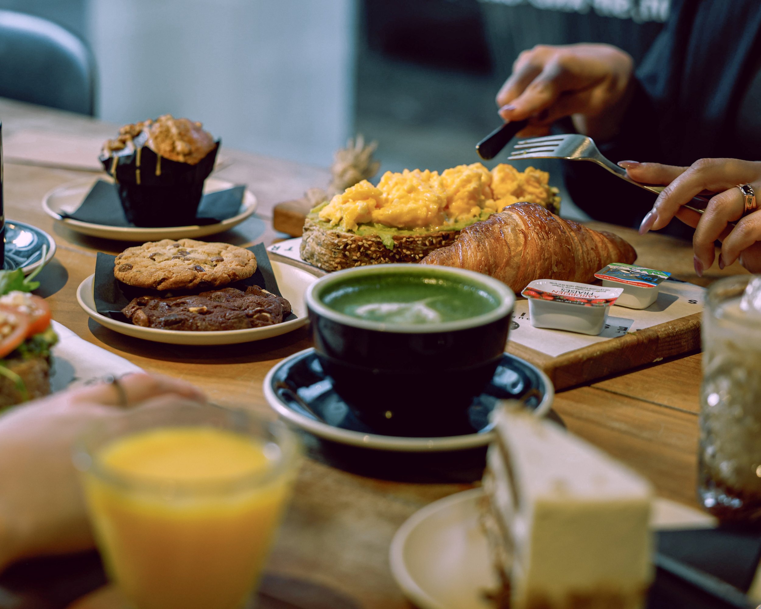 A table filled with breakfast foods including a croissant, scrambled eggs, muffins, cookies, a cup of green tea, orange juice, and small containers of cream and jam.