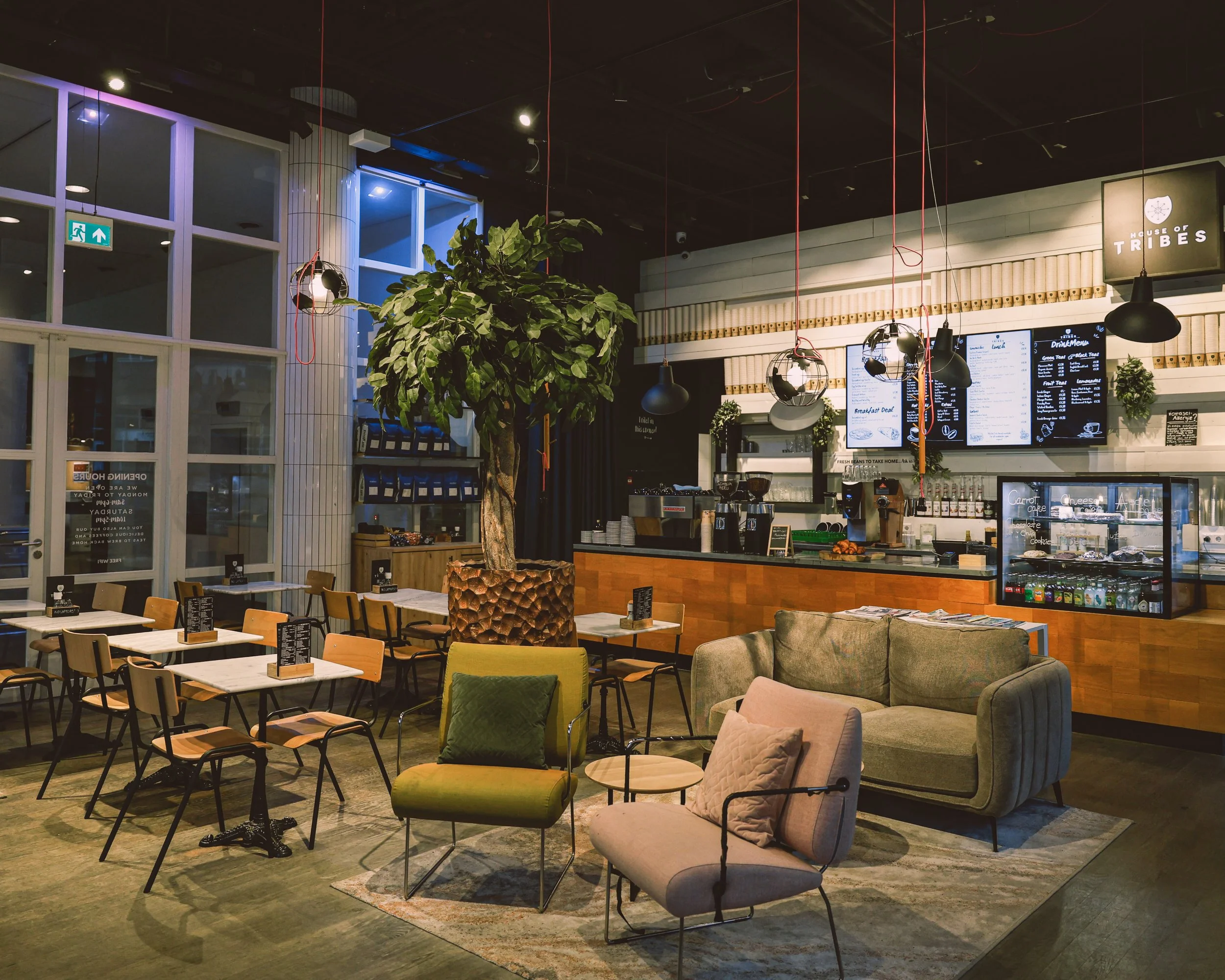 Interior of a modern cafe with a green plant in the center, seating arrangements including chairs and couches, a counter with digital menu screens, and decorative lighting.
