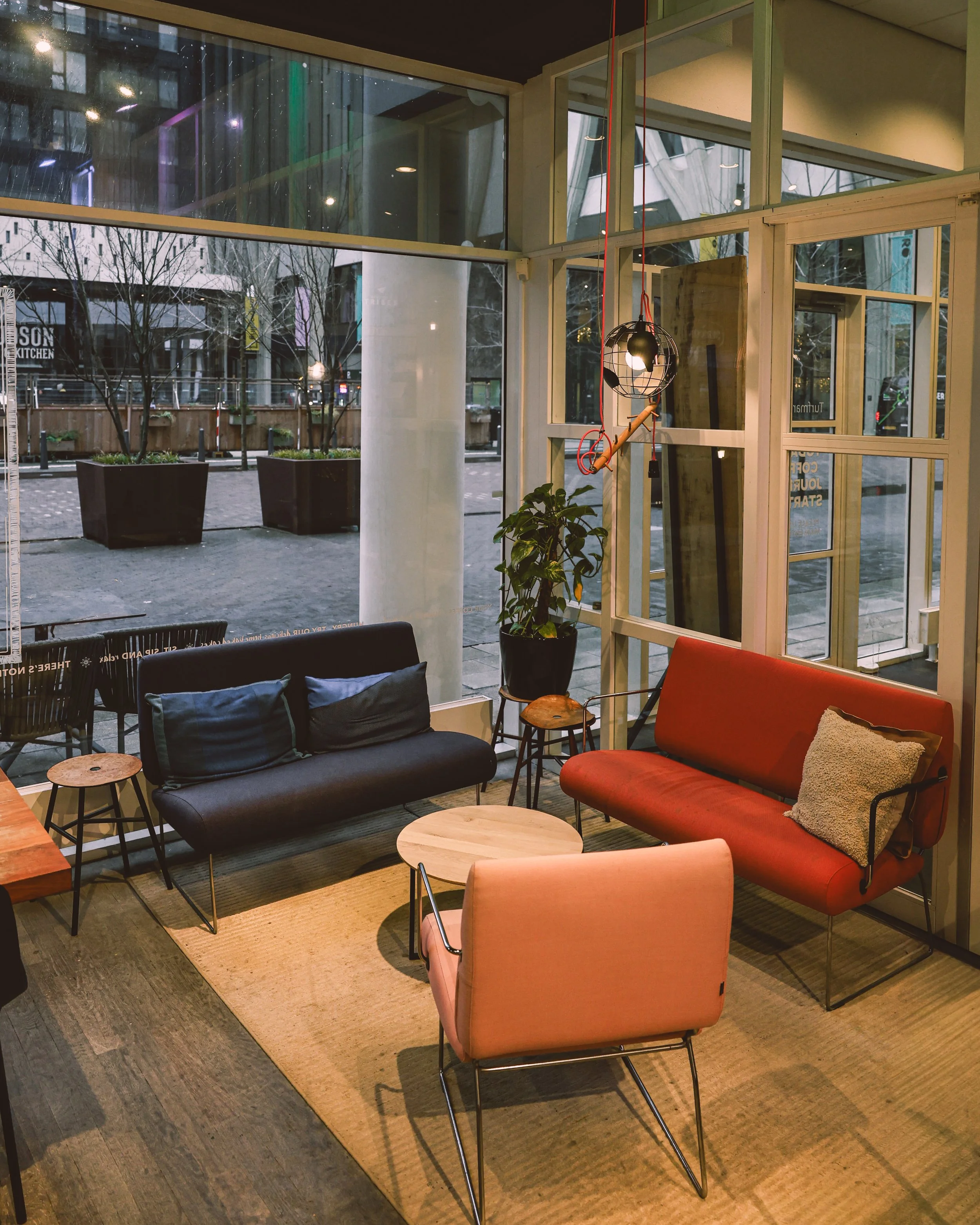 Interior of a cozy cafe with colorful modern furniture, a small round wooden table, a pink upholstered chair, a black sofa with blue pillows, a red sofa with a beige pillow, a potted plant, and large windows showing the street outside.
