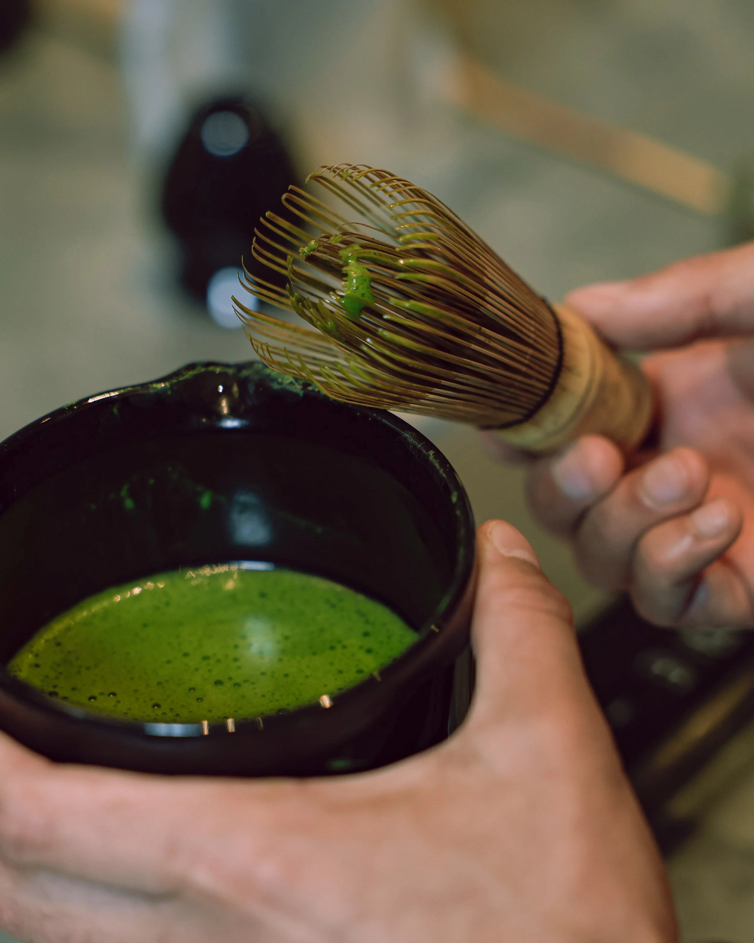 Person whisking green matcha tea with a bamboo chasen inside a black bowl.