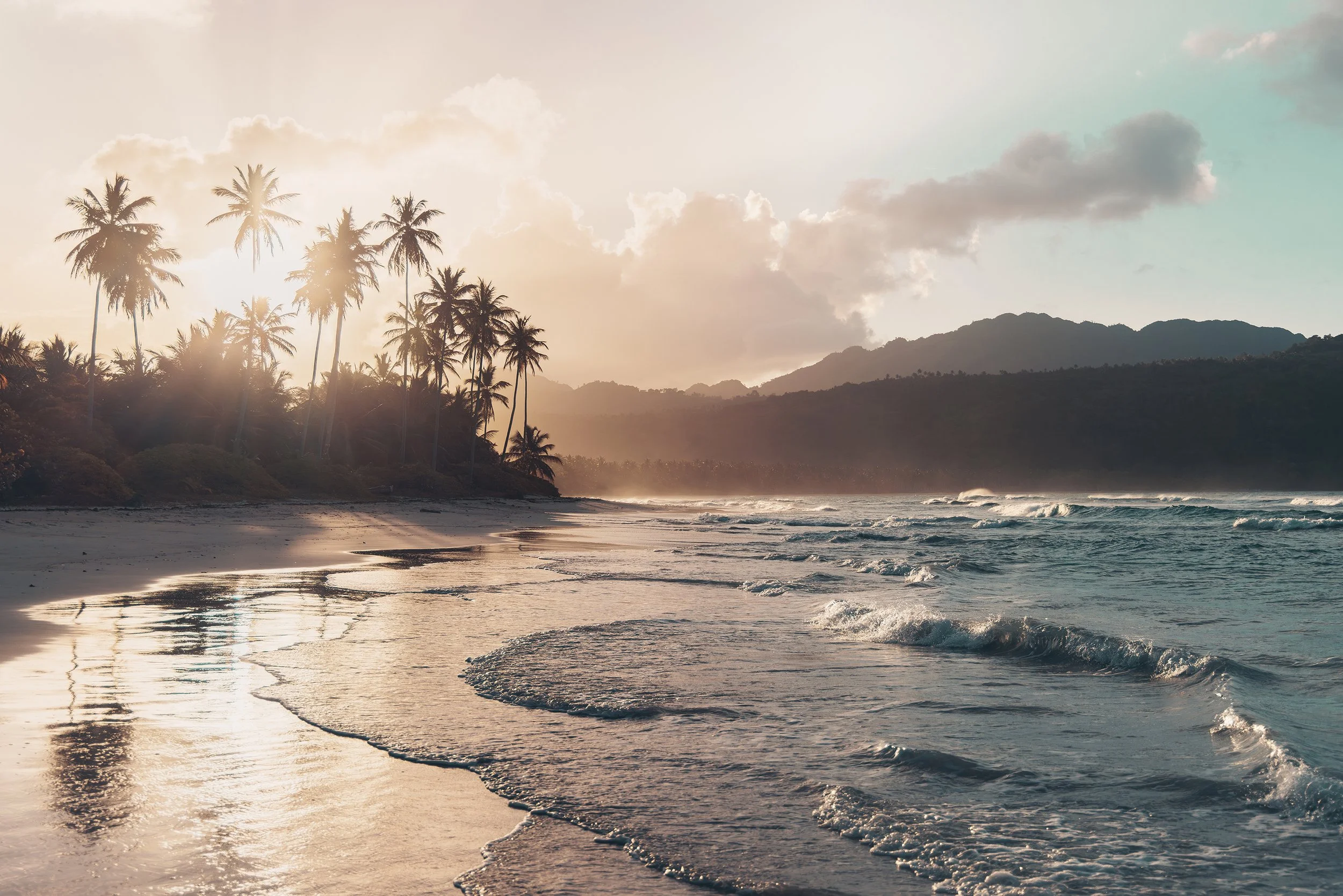 Sunset on a tropical beach with palm trees, waves, and mountains in the background.