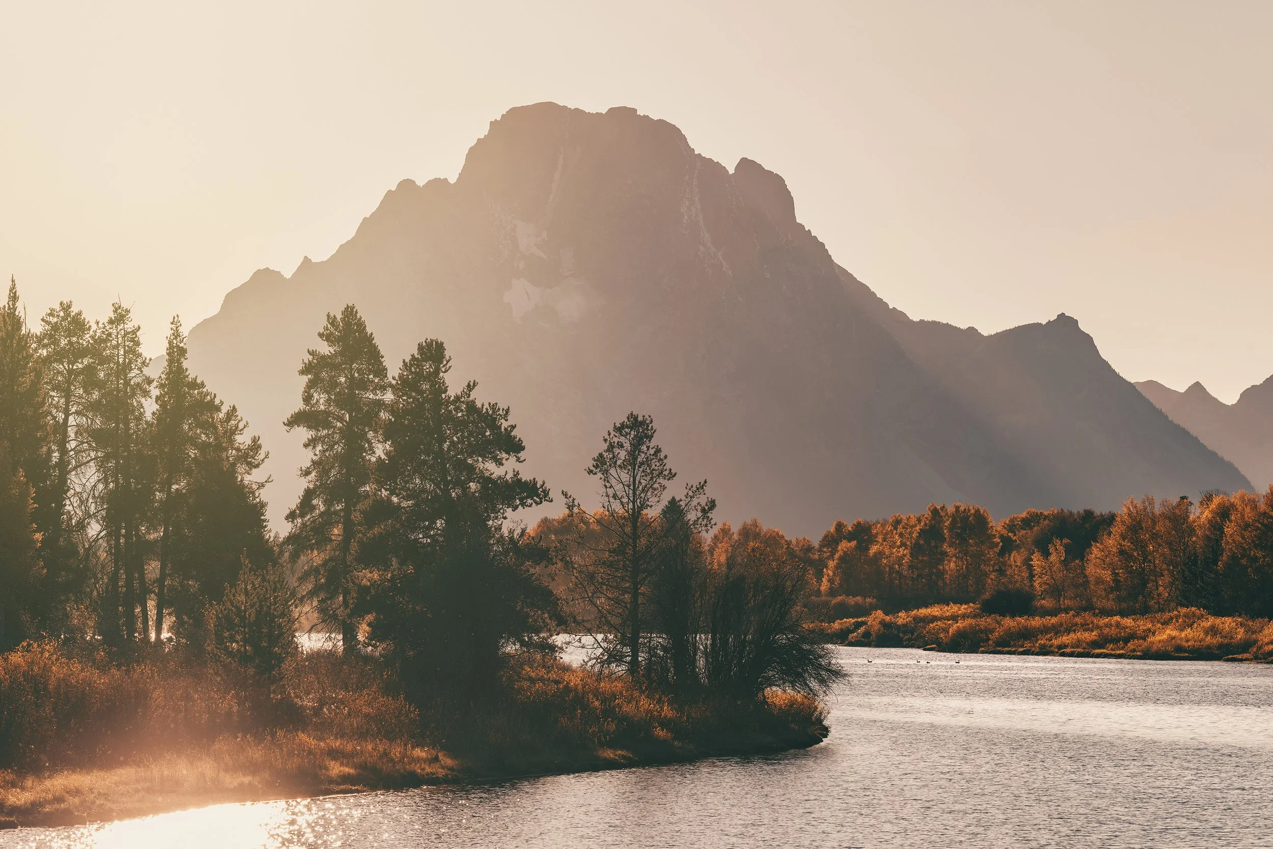 A landscape of a river flowing through a forest with trees in fall colors, and mountains in the background under a hazy sky.