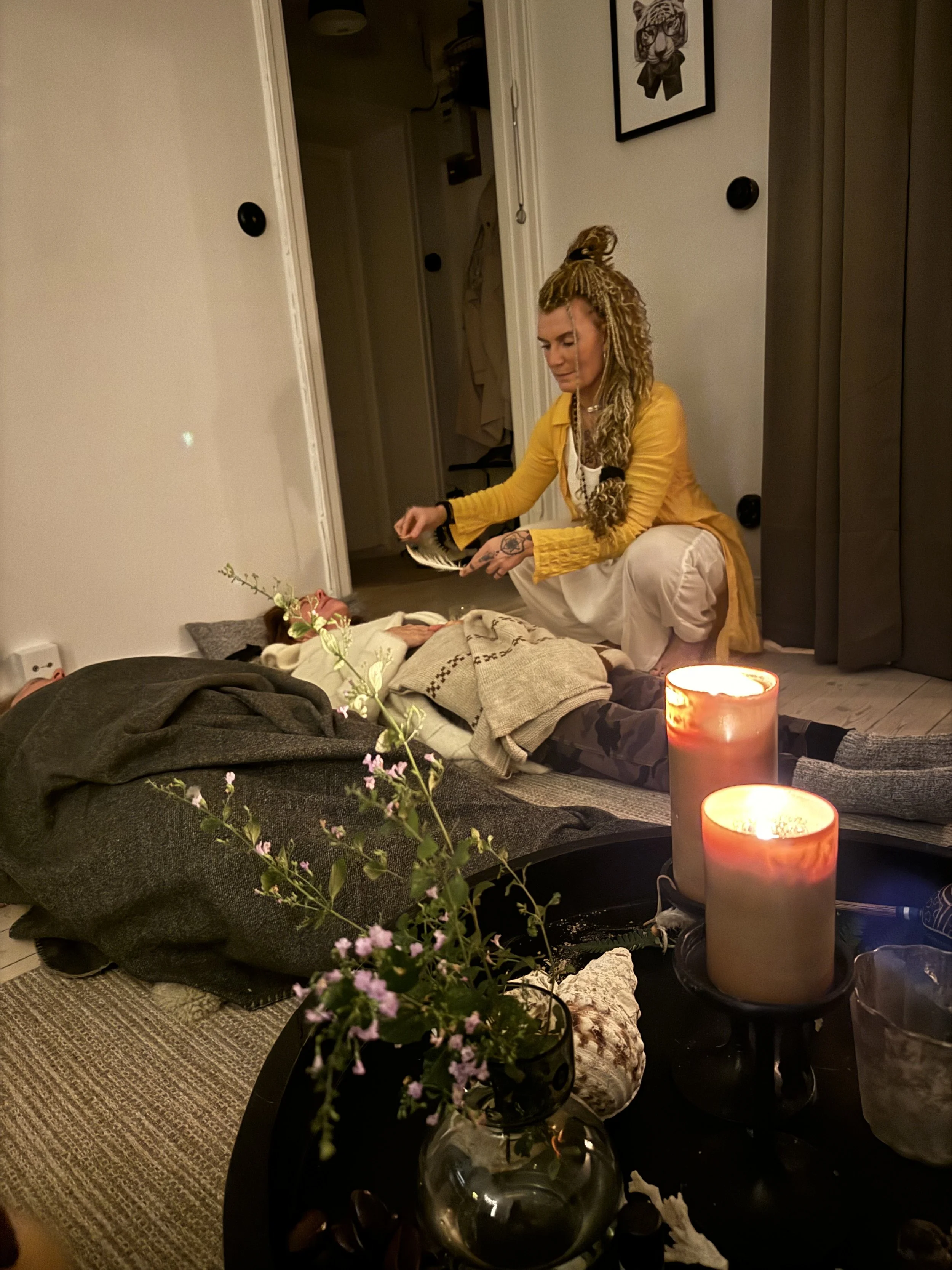 A woman kneeling next to a person lying on the floor during what appears to be a death or medical emergency, with candles and flowers nearby in a dimly lit room.