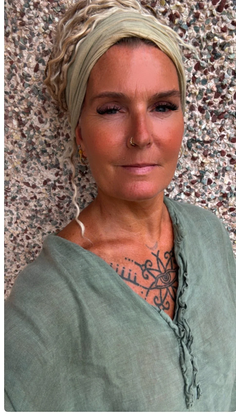 Close-up of a woman with blonde curly hair, wearing a beige headwrap, a nose ring, and earrings, lying against a textured stone wall, dressed in a green top, with visible tattoos on her chest.