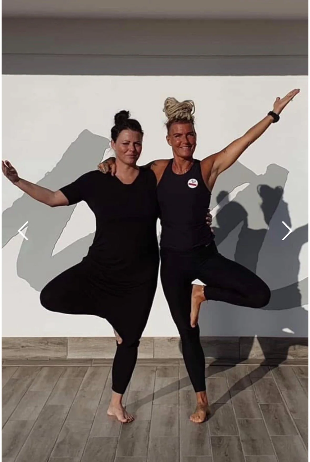 Two women practicing yoga indoors, balancing on one leg with arms outstretched, shadows cast on the wall behind them.