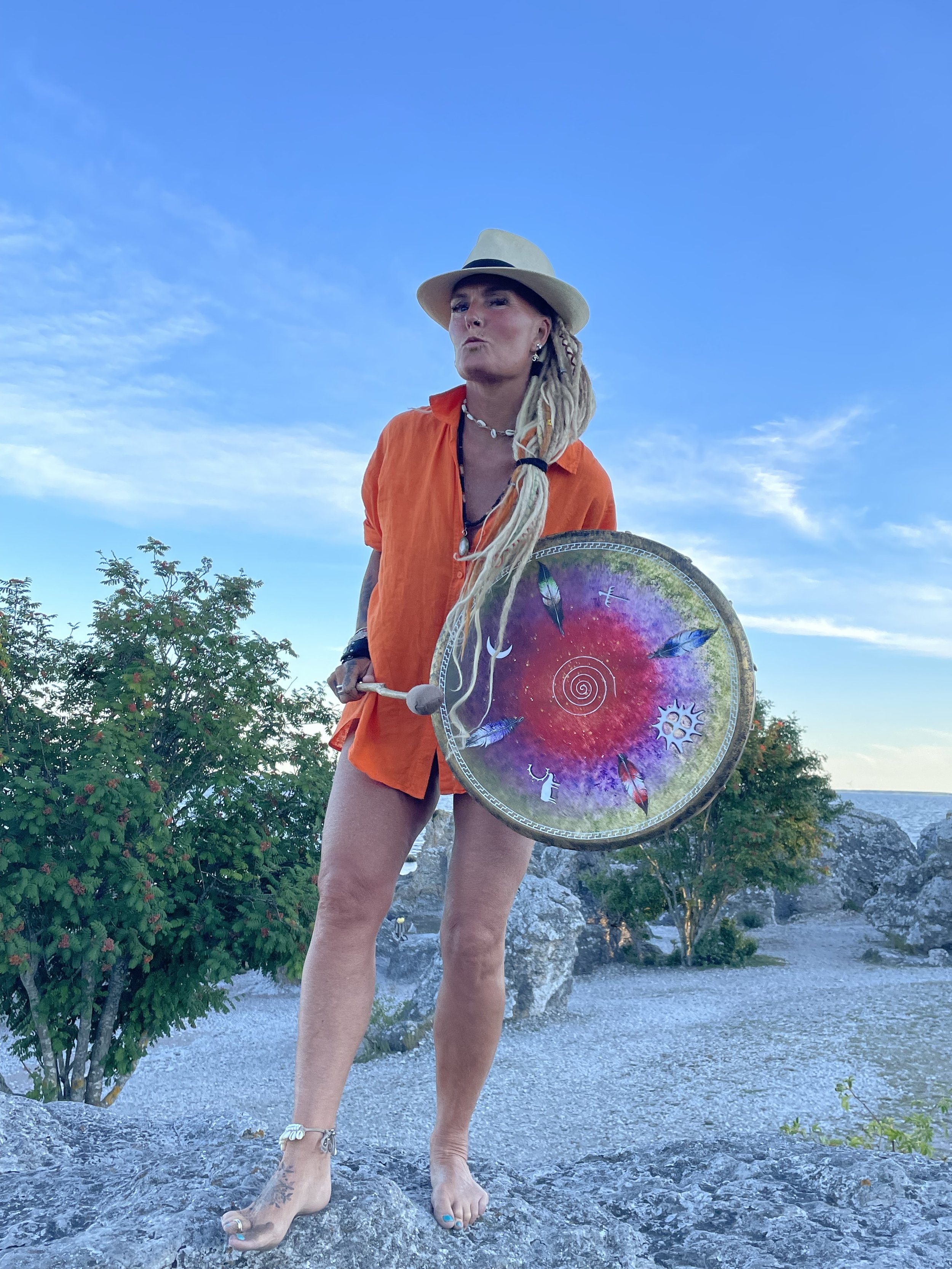 Woman with long blonde dreadlocks wearing a sunhat and orange dress, holding a colorful shield with a cosmic design, standing outdoors on rocky terrain under a blue sky with some clouds.