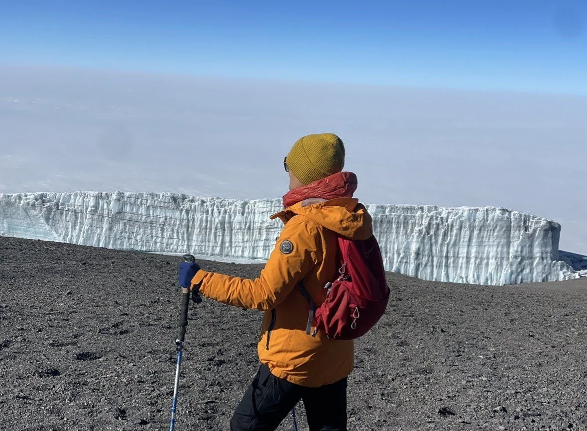 Person in an orange jacket, red backpack, yellow beanie, and gloves standing on rocky terrain, holding trekking poles, with a large ice formation and cloudy sky in the background.