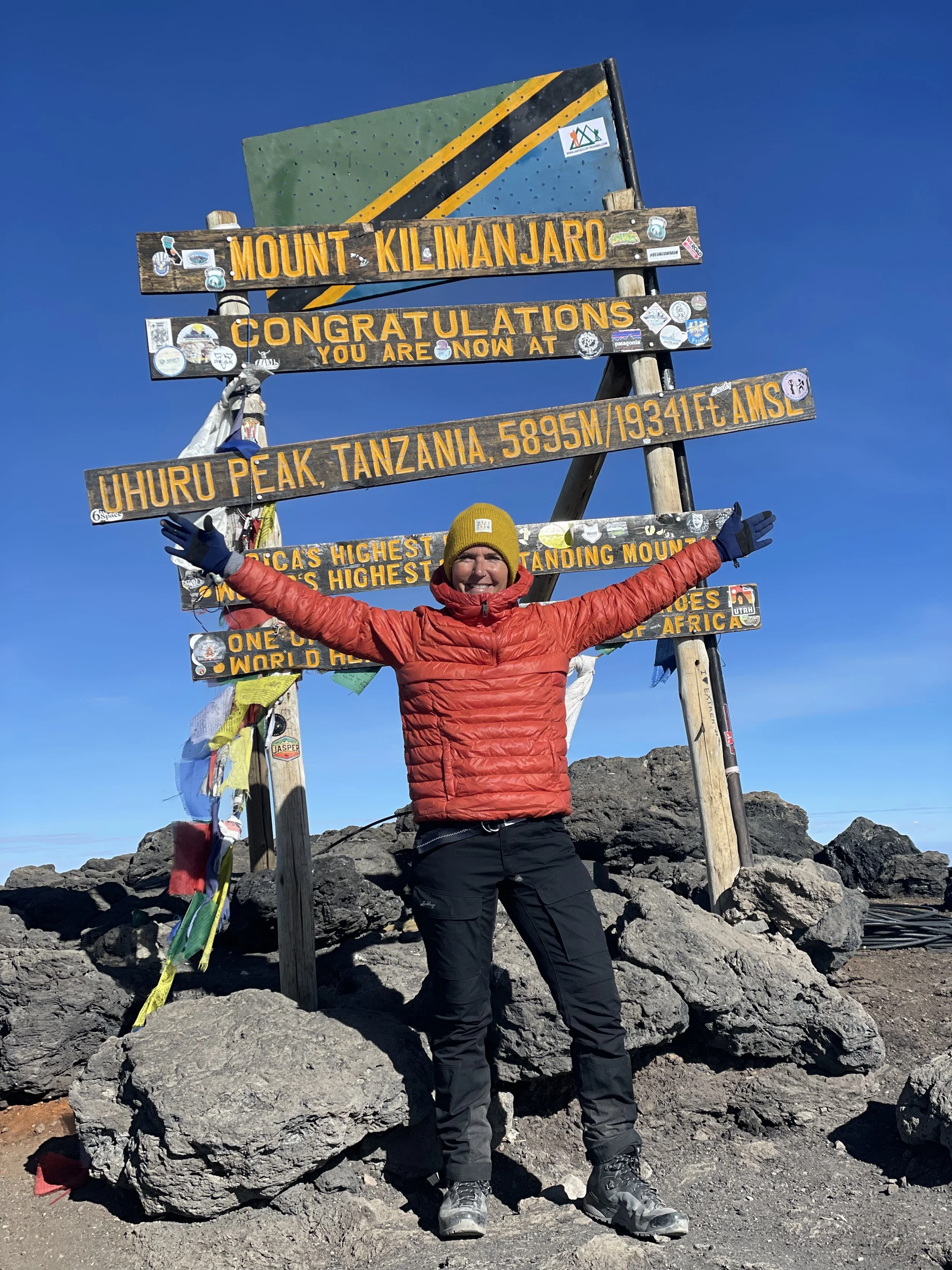 Person in a red jacket, yellow beanie, and black pants standing on rocks at the summit of Mount Kilimanjaro with arms outstretched in front of a colorful signboard marking the summit. The signboard indicates the peak's altitude of 19,341 feet and features various stickers.