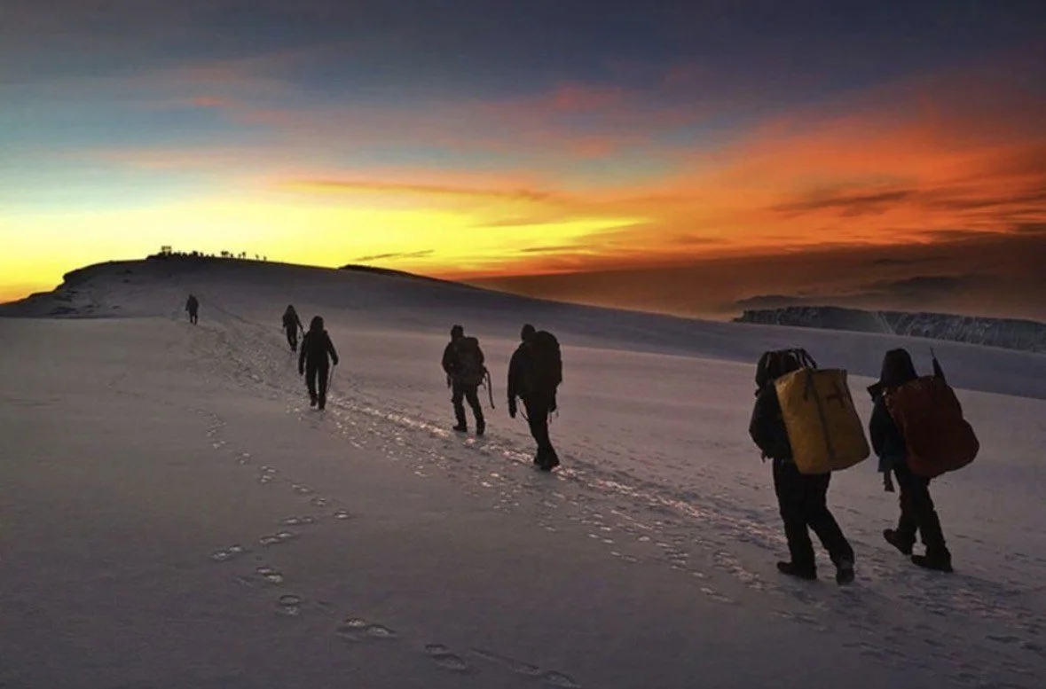 Group of hikers walking in snow during sunset with colorful sky.