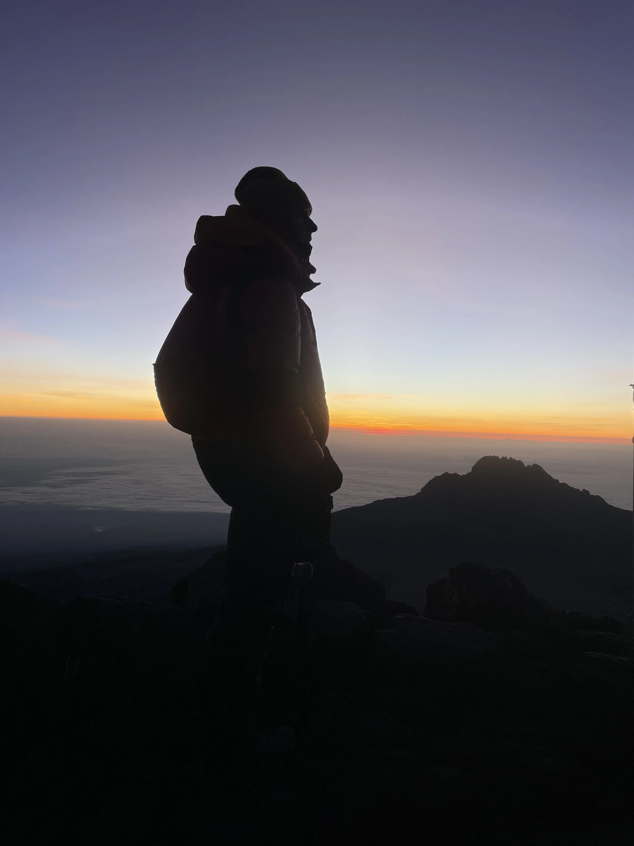 Silhouette of a person standing on a mountain at sunset with a colorful sky above and distant mountain peaks.