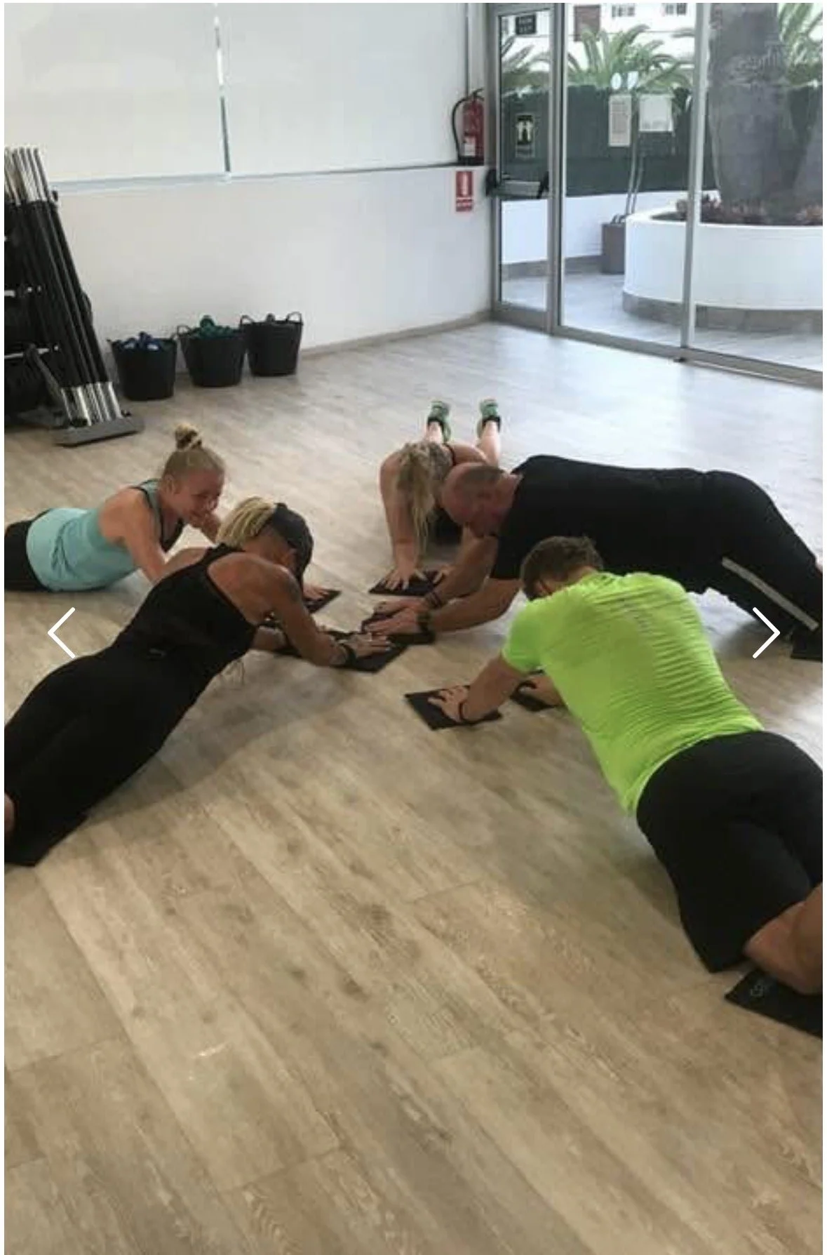 People participating in a Pilates class on mats, in a room with light wood flooring, near a large glass window and doors, some lifting their upper bodies and others stretching on their stomachs.