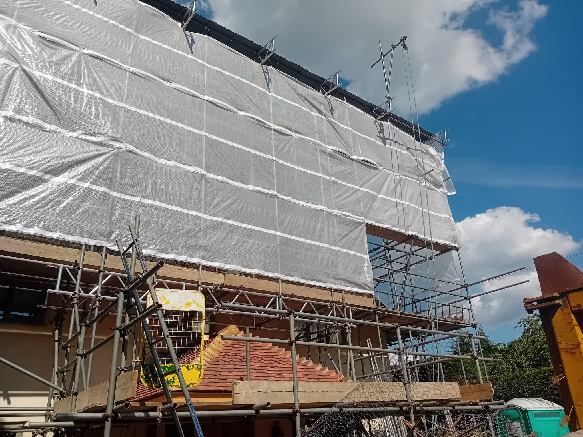 Building under construction with scaffolding and white protective sheeting, a roof with red tiles, a portable toilet, and a cloudy sky.