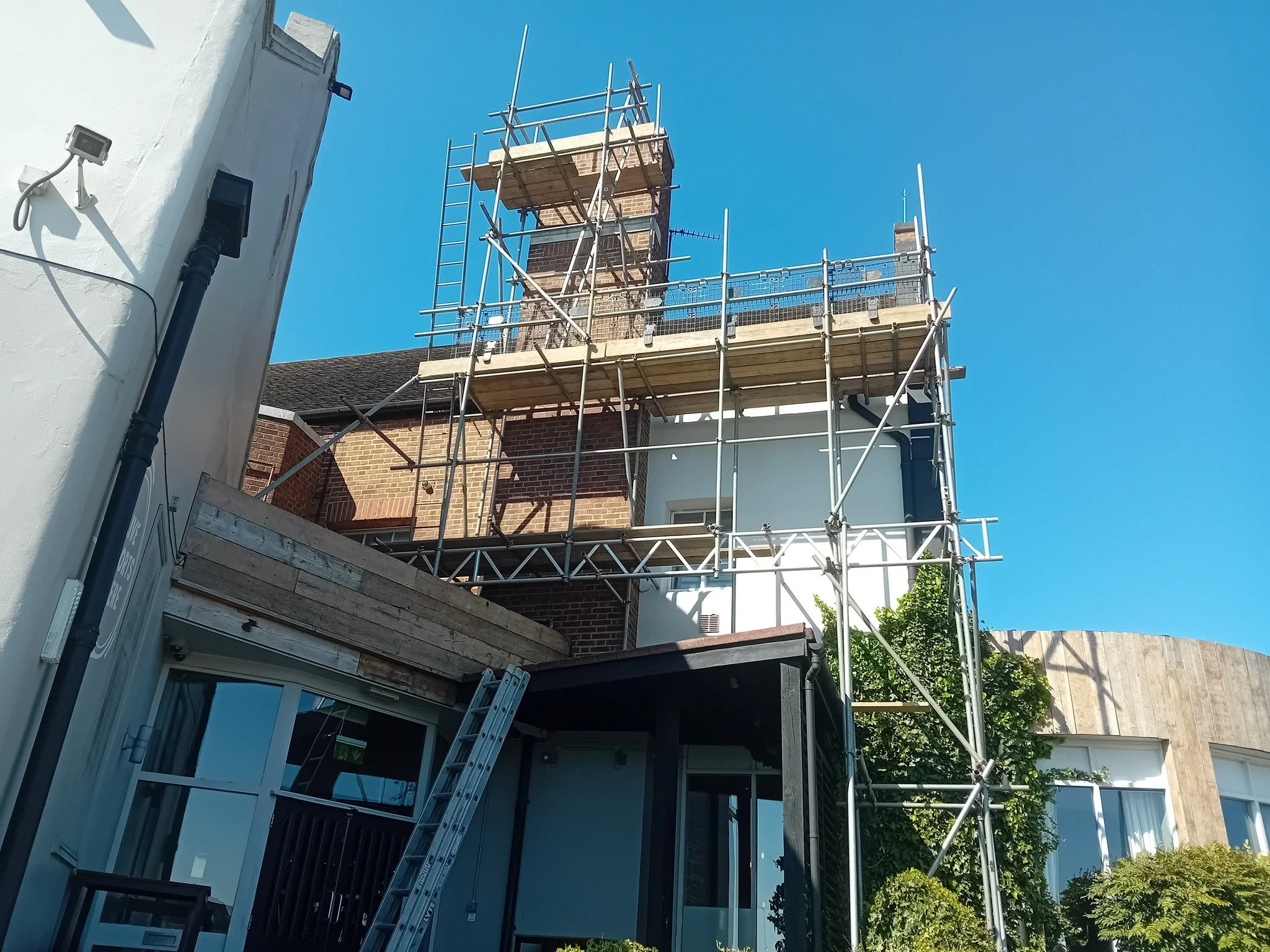 Building under construction with scaffolding on the upper floors, blue sky in background, and some greenery.