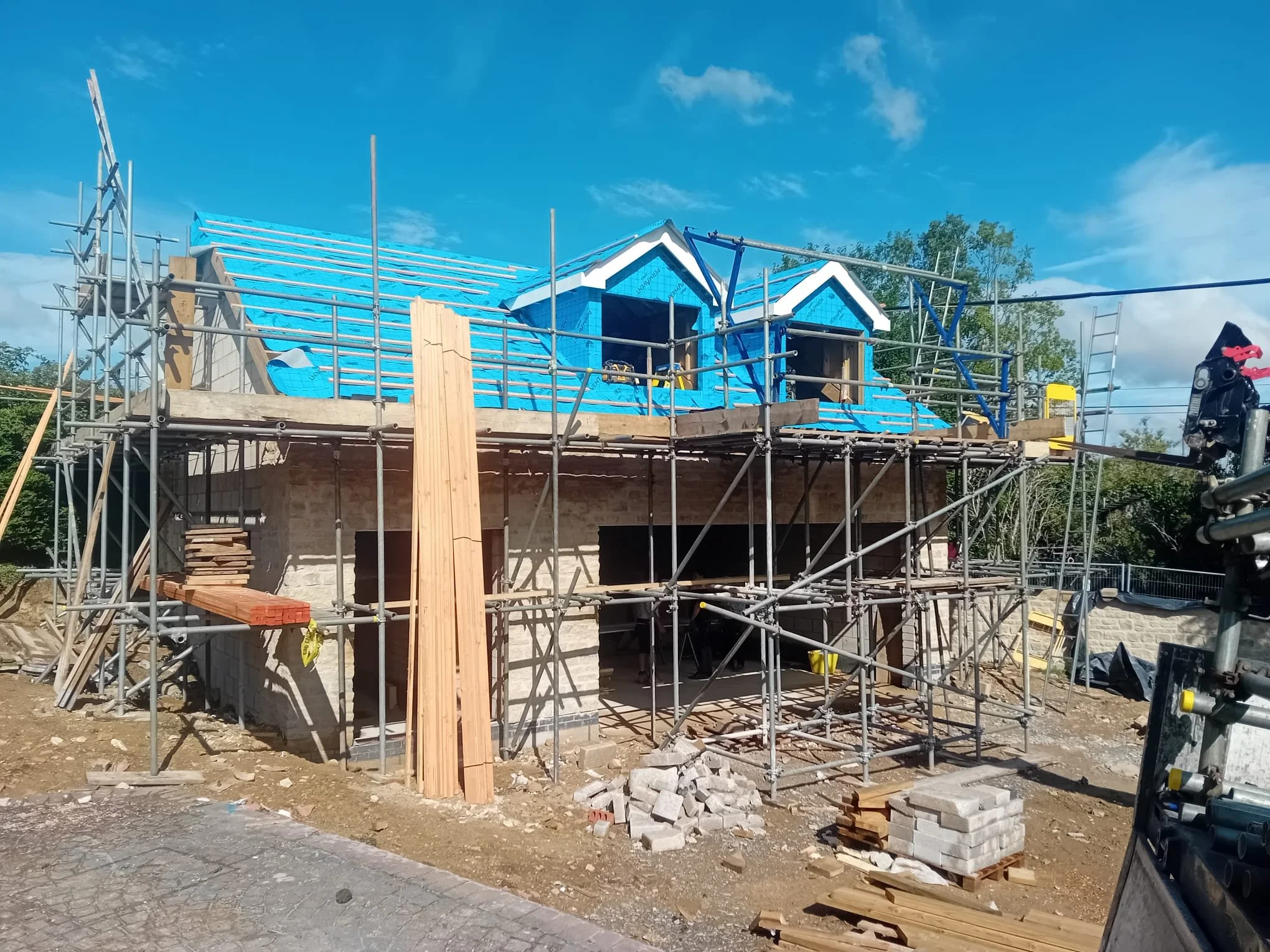 A house under construction with scaffolding around it, and blue roofing materials installed on the roof, with some windows framed but not yet fitted.