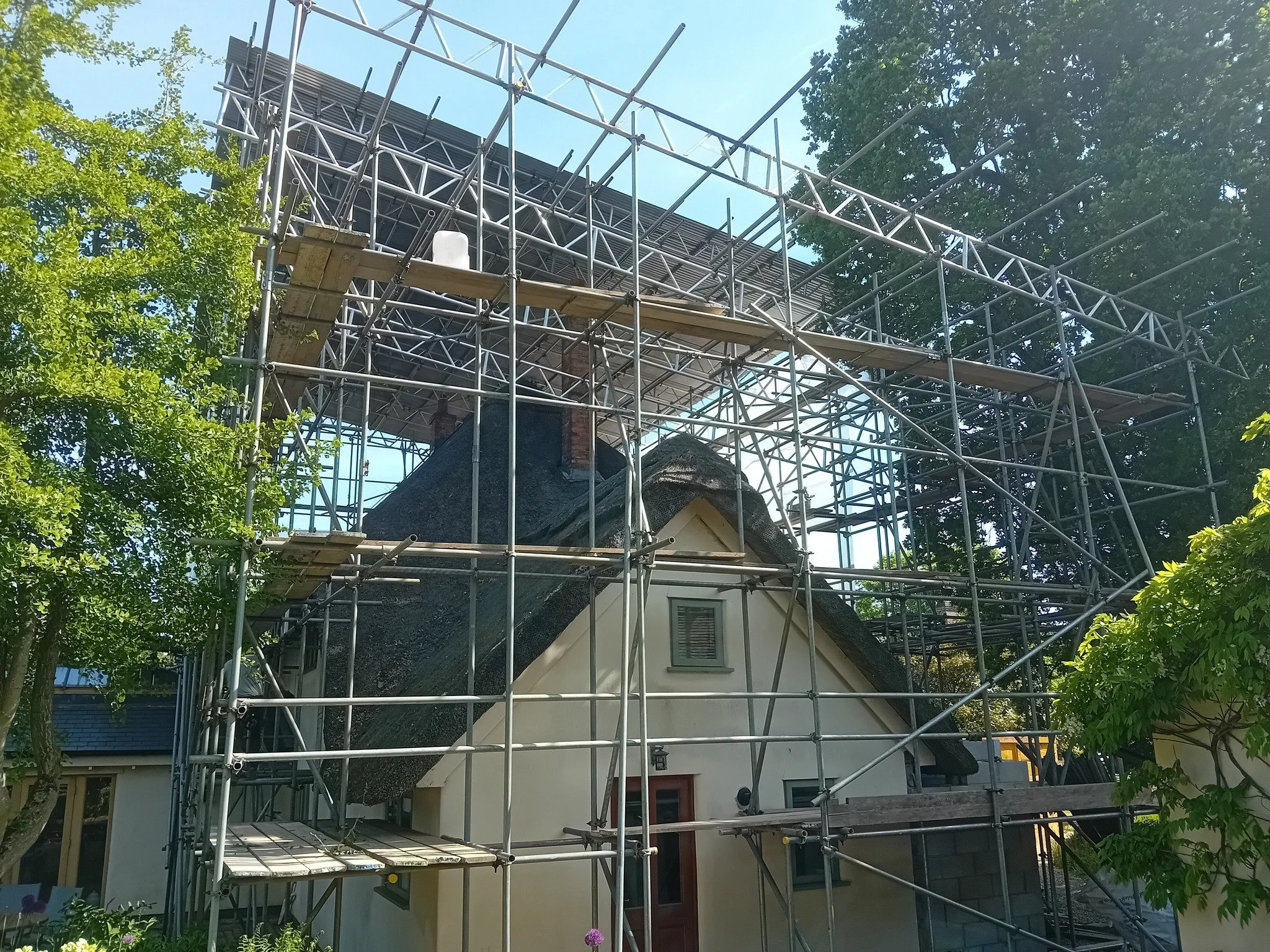 A house with a thatched roof surrounded by scaffolding during construction or renovation, with green trees and blue sky in the background.