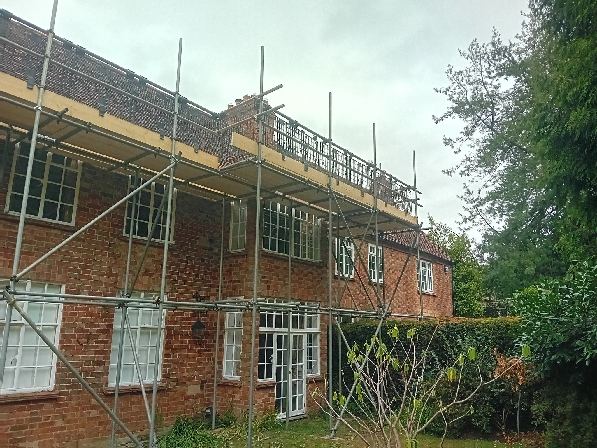 A two-story brick house with white window frames, a glass door, and a chimney, surrounded by greenery. Scaffolding is set up around the upper part of the house for construction or maintenance work.