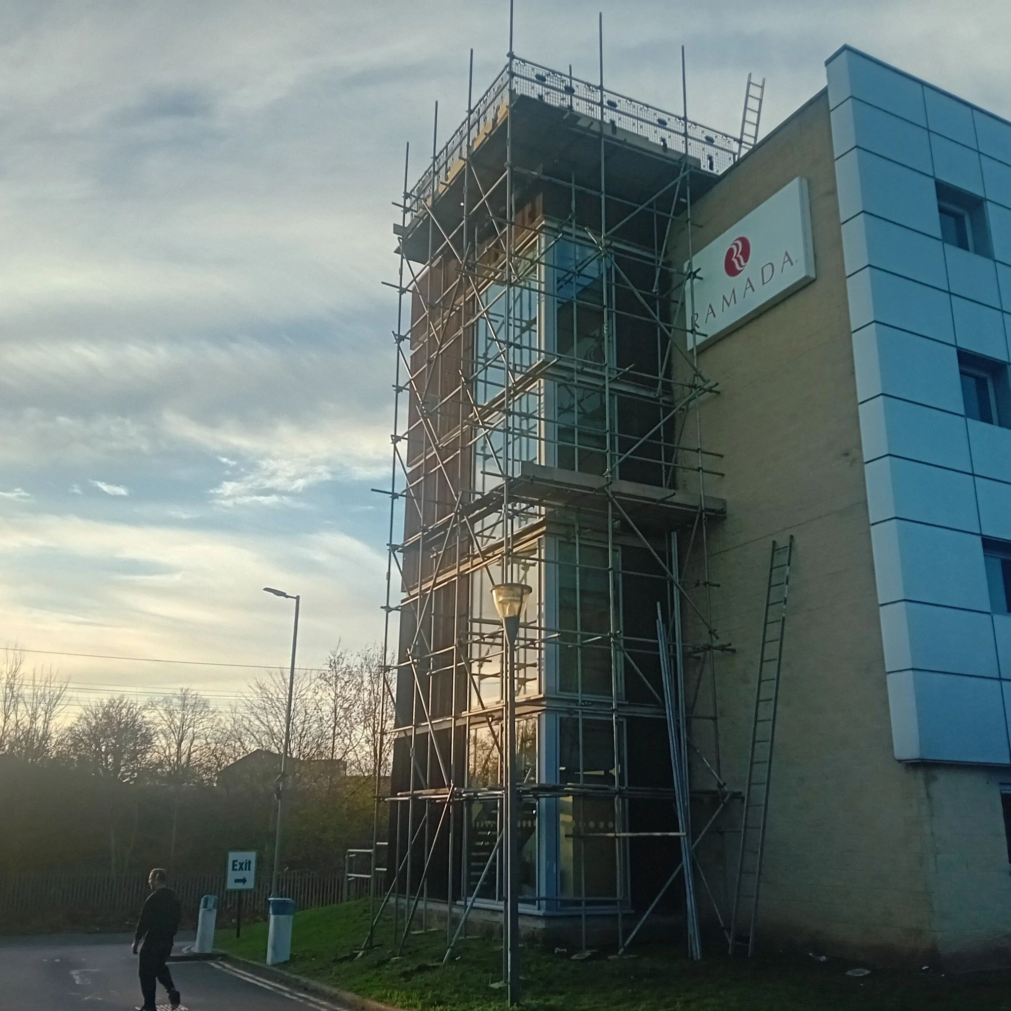A building with scaffolding on the front, with a sign that reads 'RAMADA'. A person is walking on the sidewalk near the building, and there is a streetlamp and an overhead street sign that says 'Exit'. The sky is partly cloudy and the time appears to be late evening or early morning.