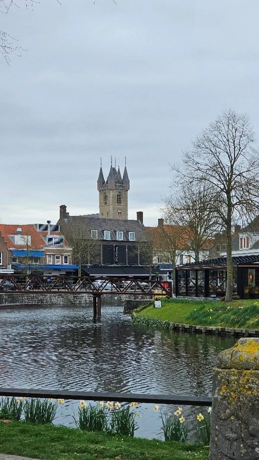 Een gezicht op een rivier met een brug, omringd door gebouwen en een kerk met torenspitsen in de achtergrond, tijdens bewolkt weer.
