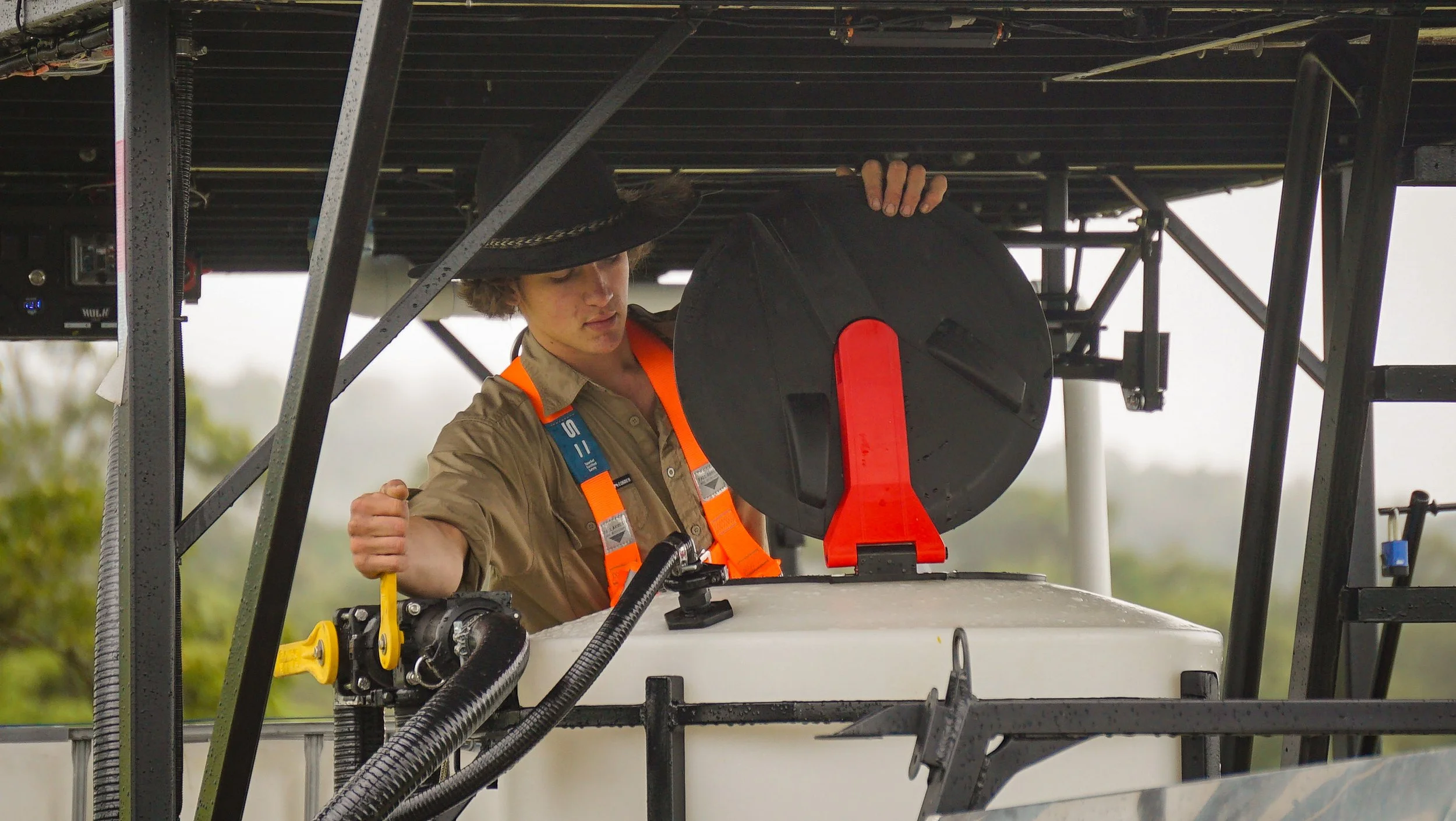 A person wearing a scapular hat and khaki shirt working on a mechanical or aircraft component under a metal framework.