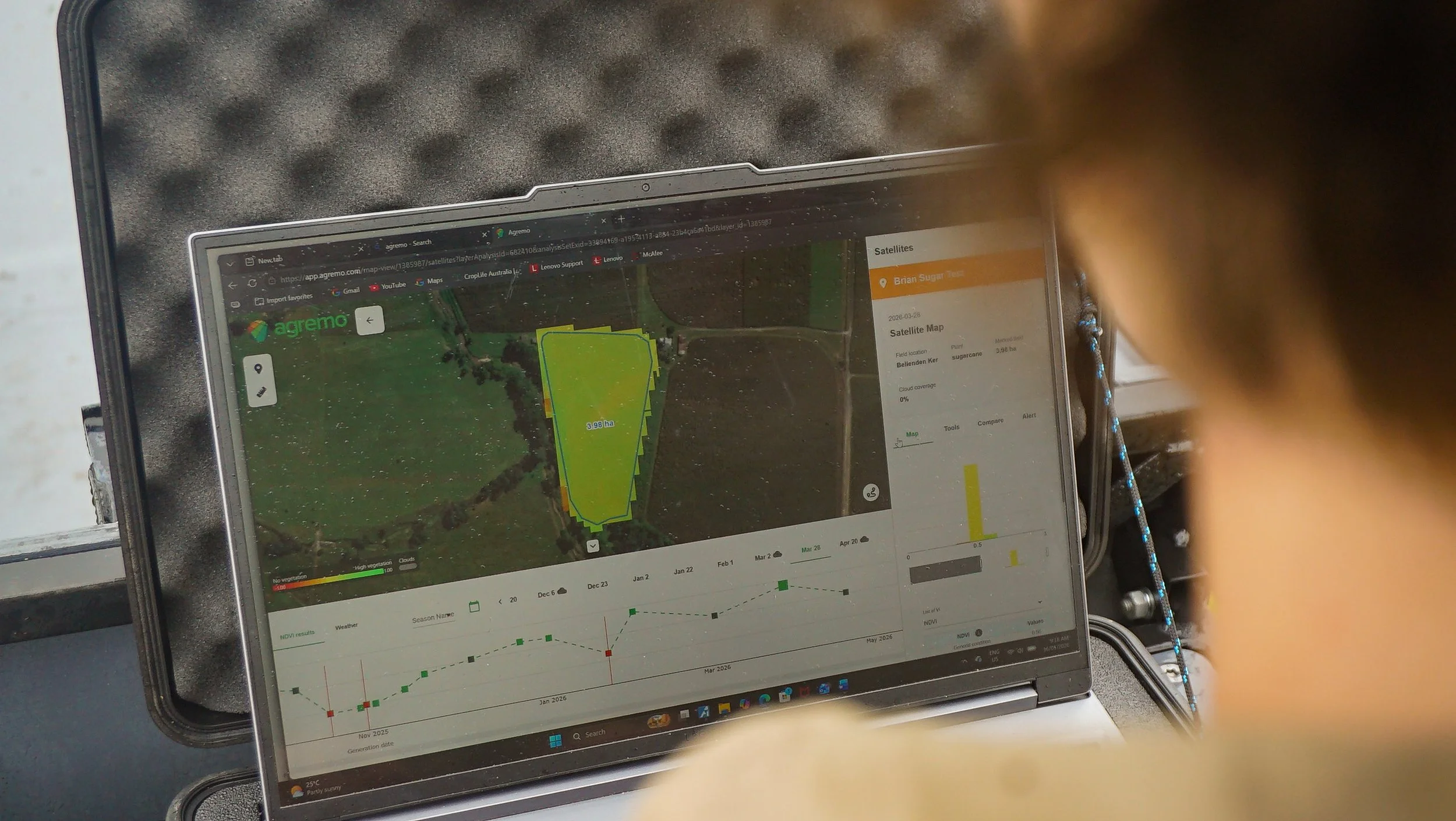 A laptop on a gray cushioned surface displays a satellite map and data analytics related to field management, with a person partially visible and blurred in the foreground.