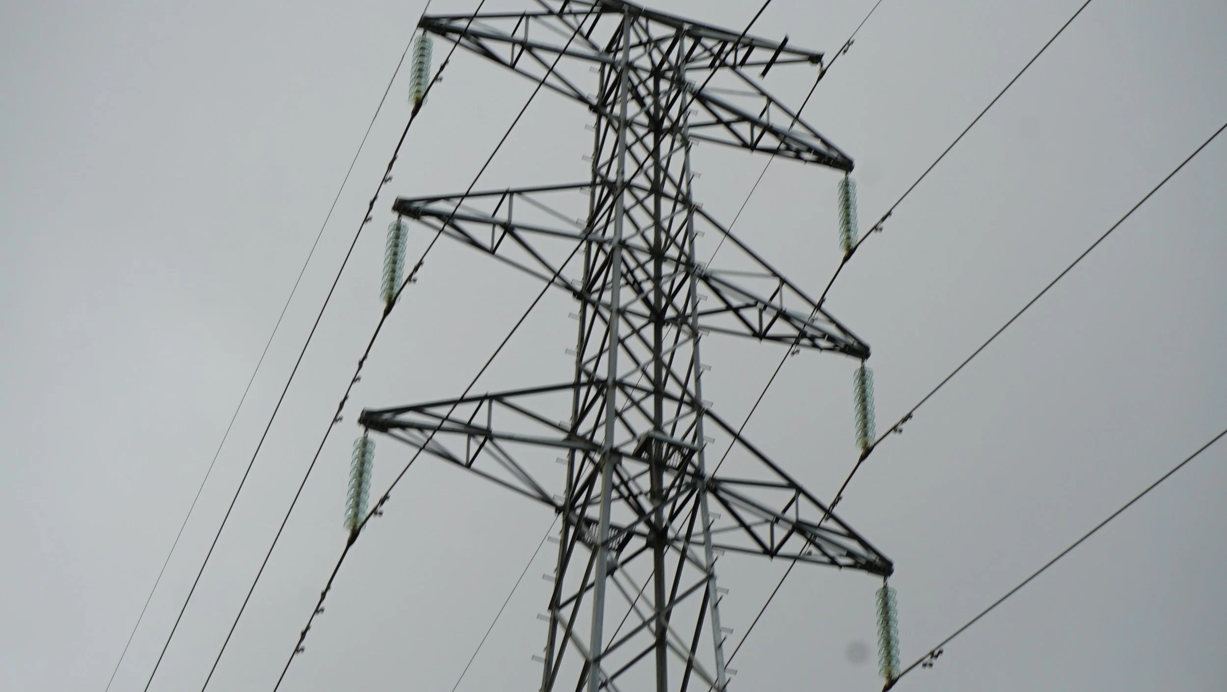 A close-up view of an electrical transmission tower with power lines against a gray overcast sky.